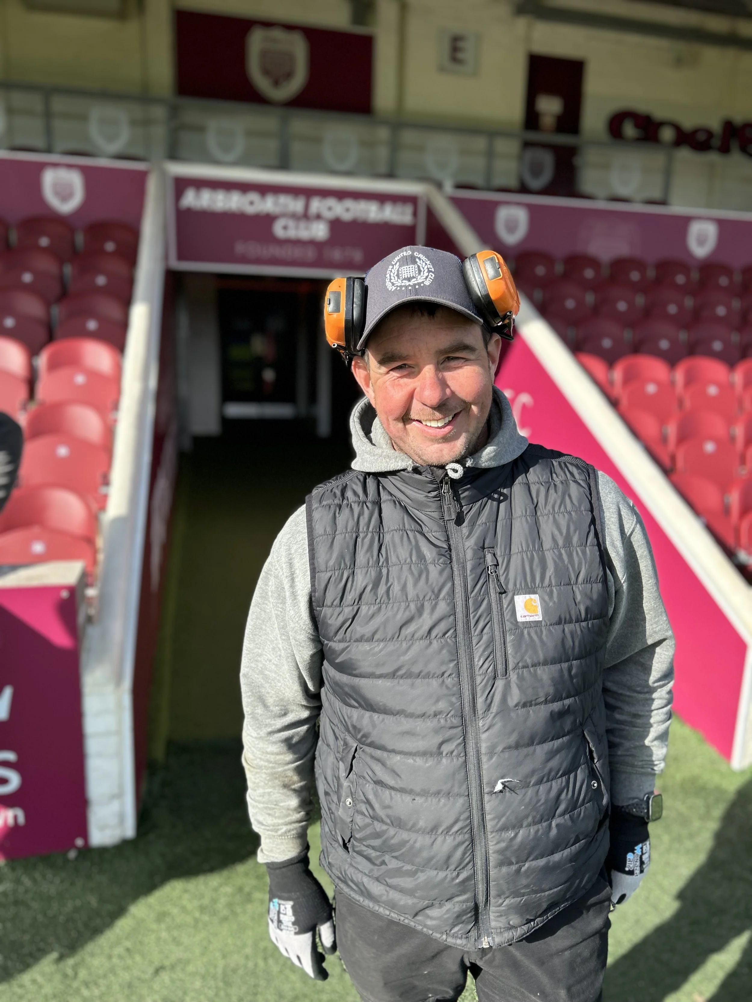 Todd Smith author, smiling at a football stadium, wearing a black Carhartt vest, gray hoodie, and protective ear muffs with the Auburn Football Club sign in the background.