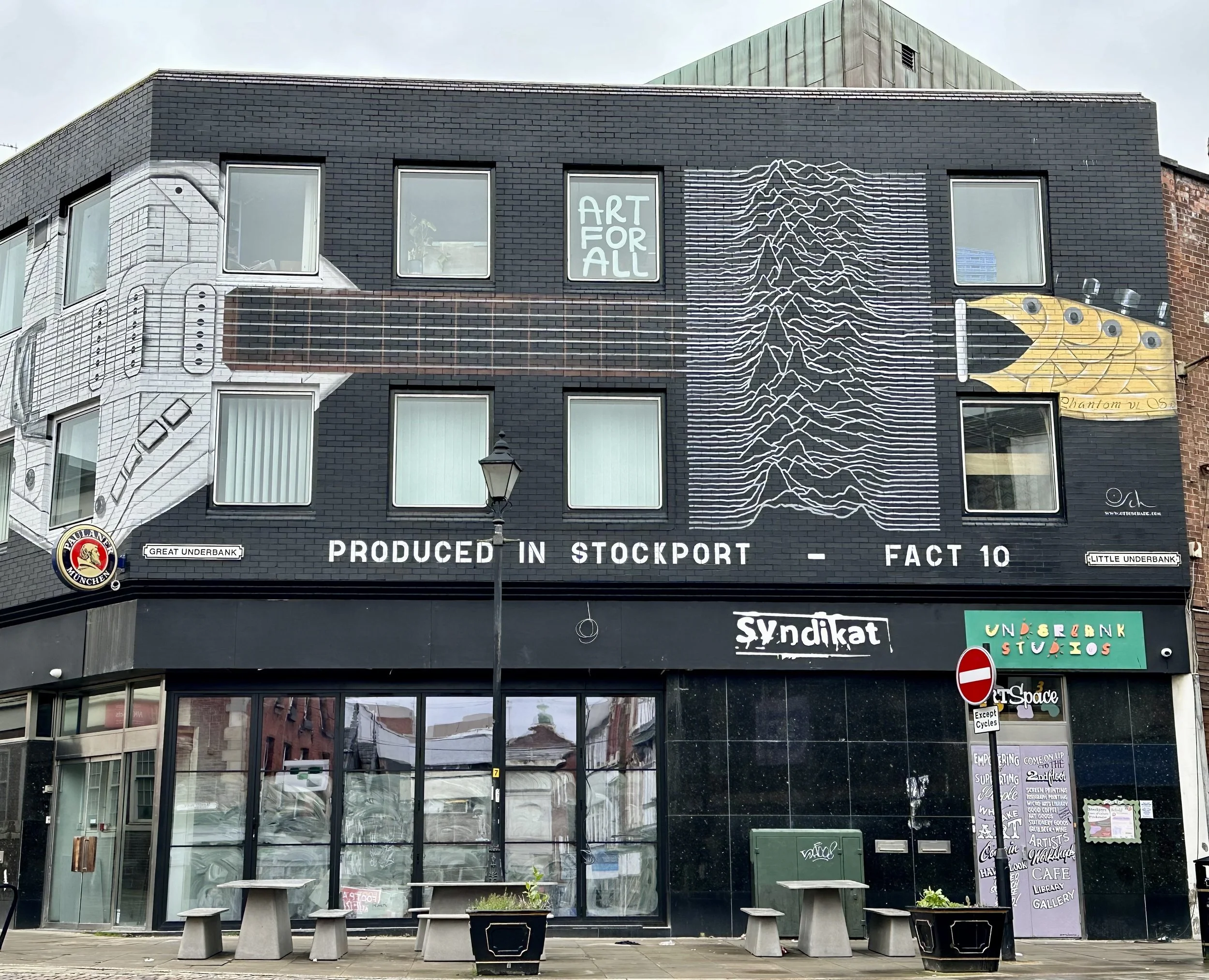 A mural painting on the black brick building depicts a white electric guitar on the left and a yellow electric guitar on the right, with lines and patterns connecting them. The mural includes text such as 'ART FOR ALL' in a window, and 'PRODUCED IN S