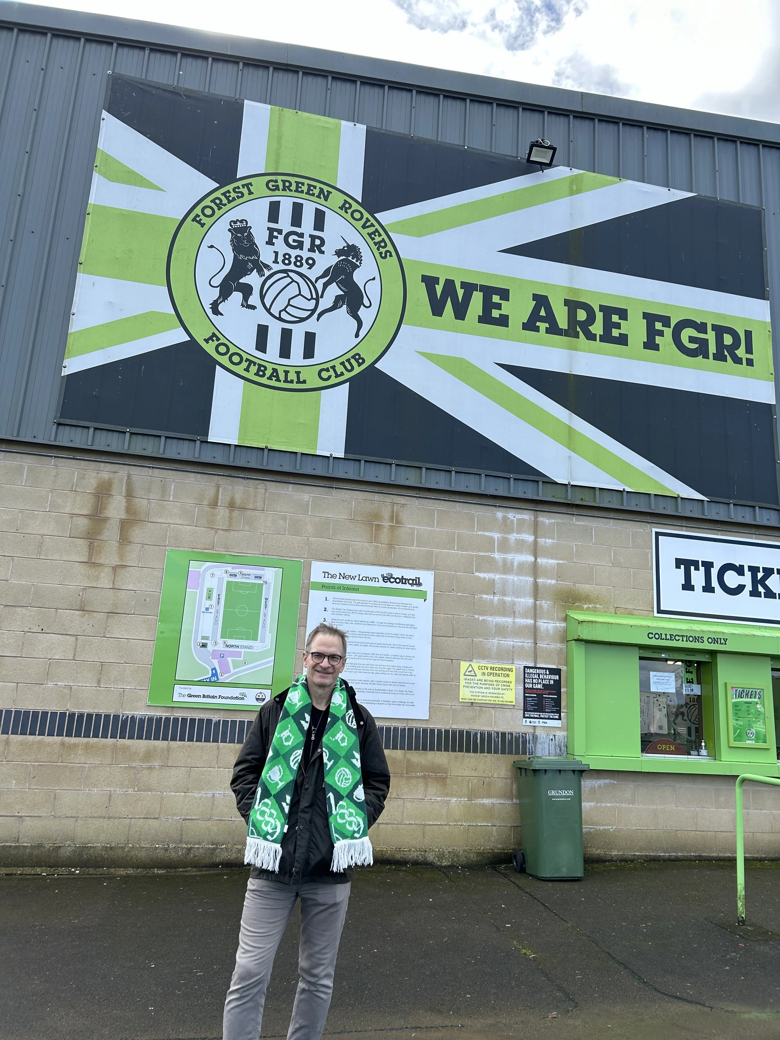 A man standing in front of a sports stadium or football ground, wearing a green and white scarf, with a smiling expression.