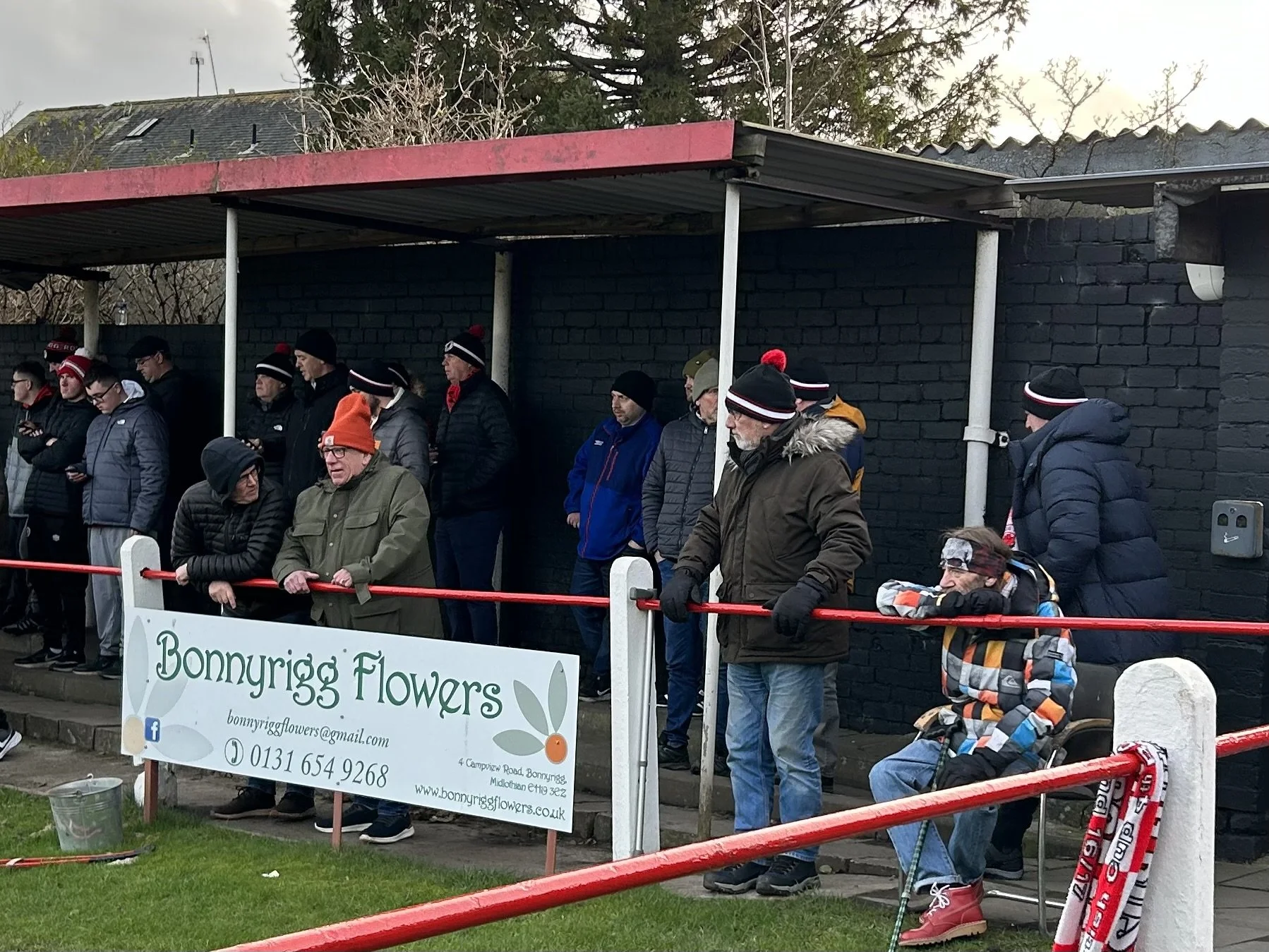 People standing in a queue outdoors, many wearing winter hats and jackets, waiting near a black brick wall with a red roof. A woman with a walker sits at the front of the line, wearing sunglasses and a colorful jacket. A sign for Bonnyrigg Flowers is