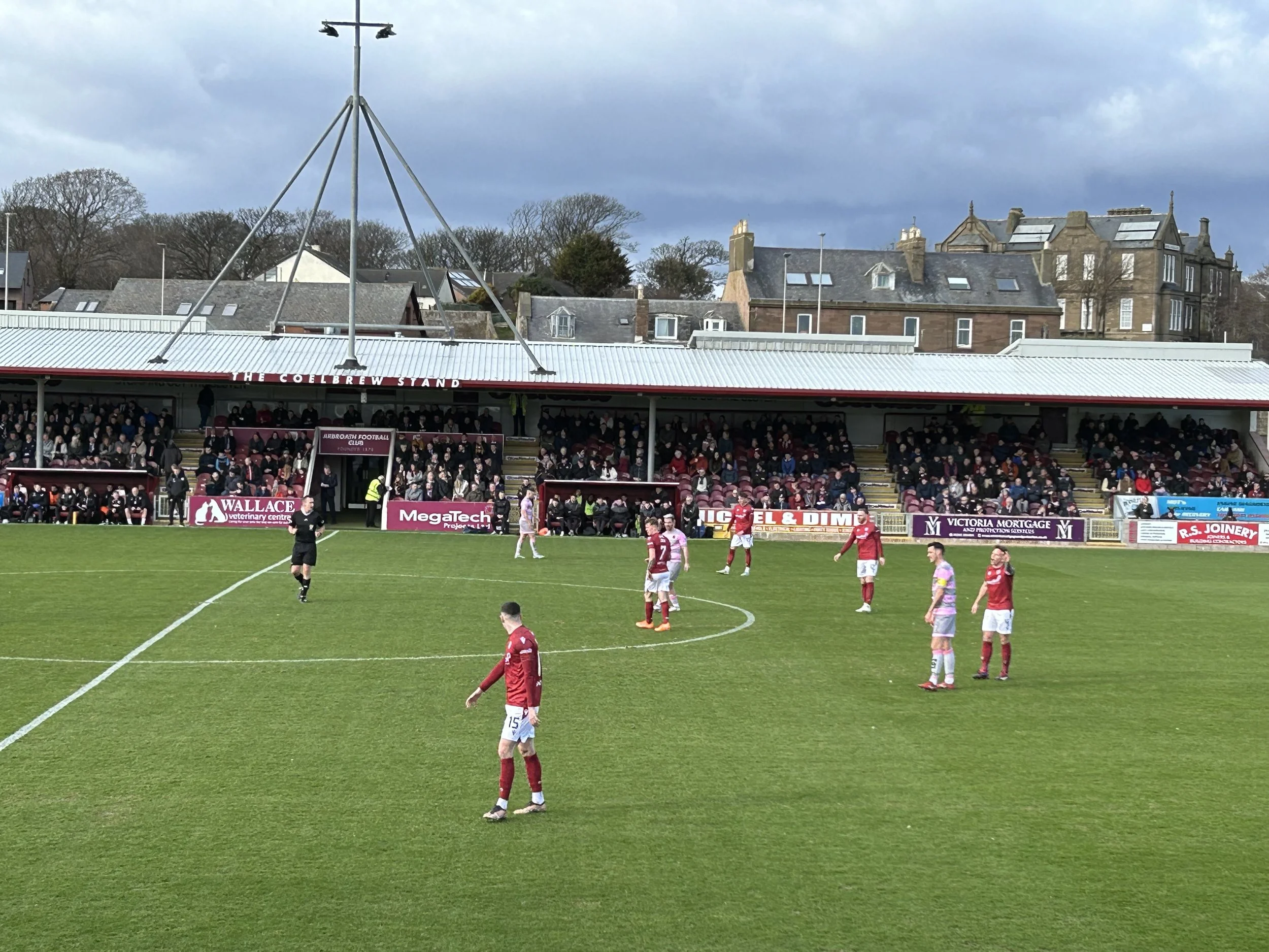Soccer match in progress with players on the field and spectators in the stands at the Coelbren Stand, with residential buildings in the background under a cloudy sky.