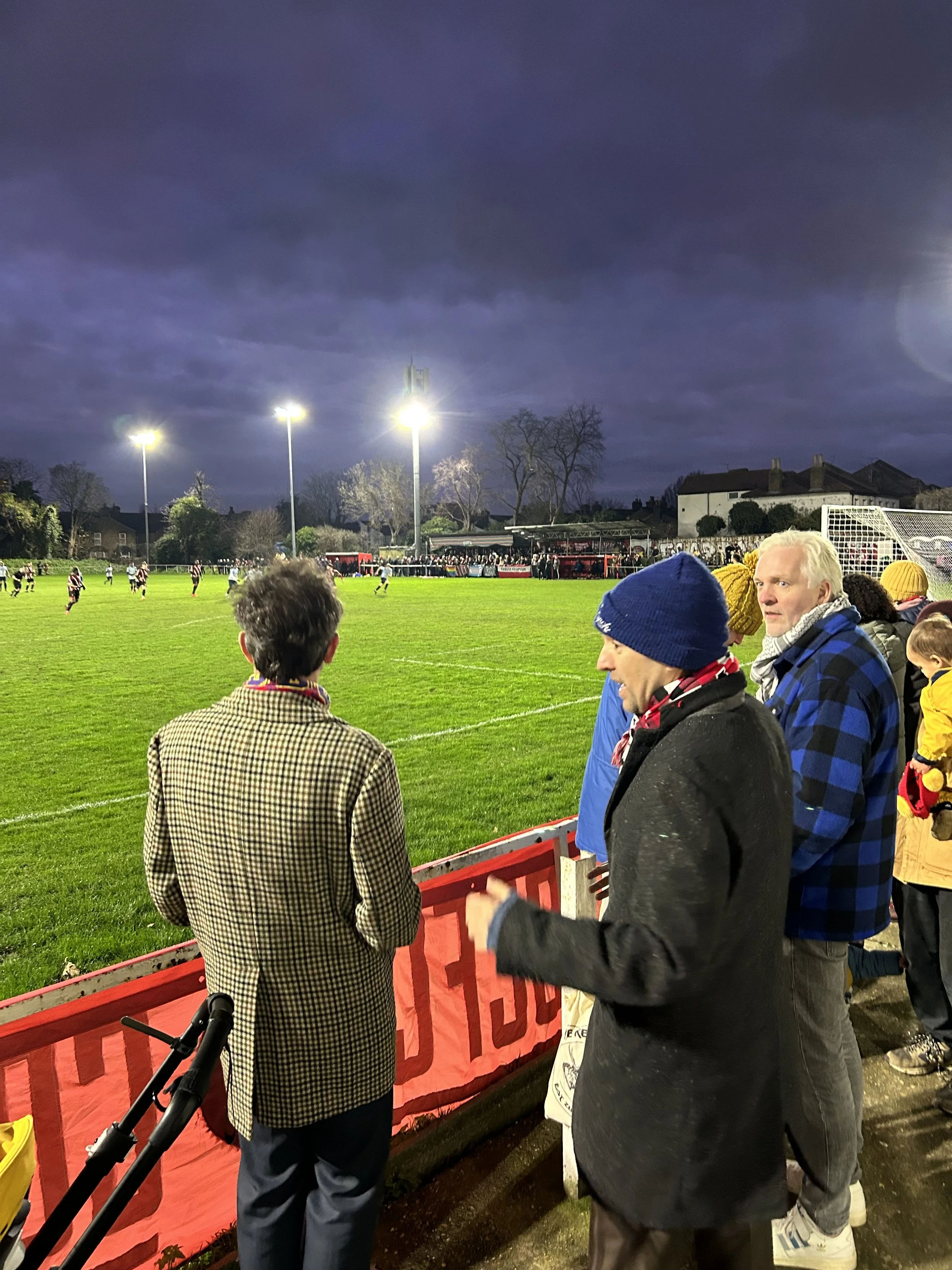 People watching a soccer game on a well-lit field in the evening, with spectators standing behind a red barrier, some wearing winter clothing.