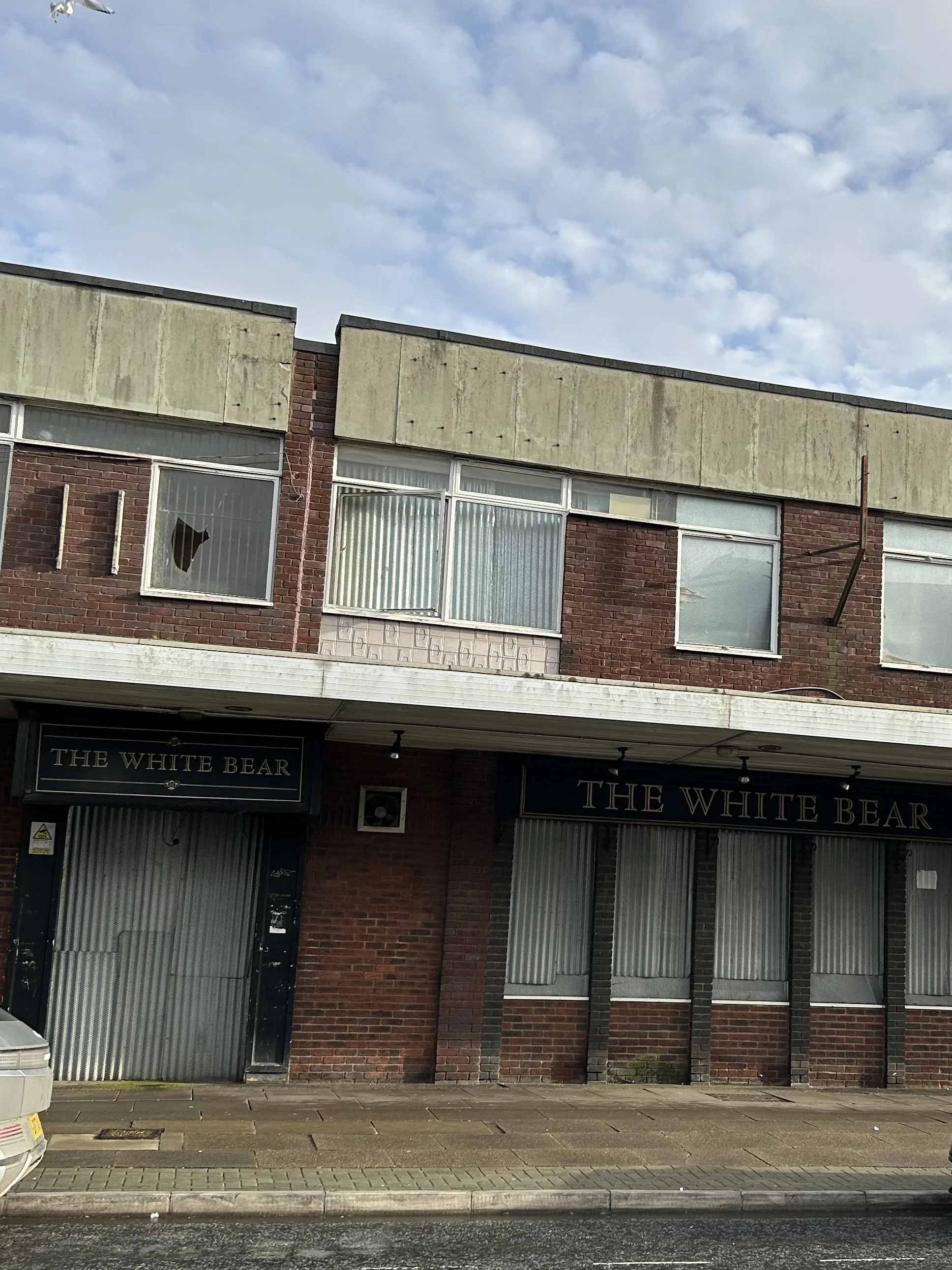 Facade of a brick building with large windows and a sign that reads 'The White Bear' above the entrance, with a broken window on the upper left side.