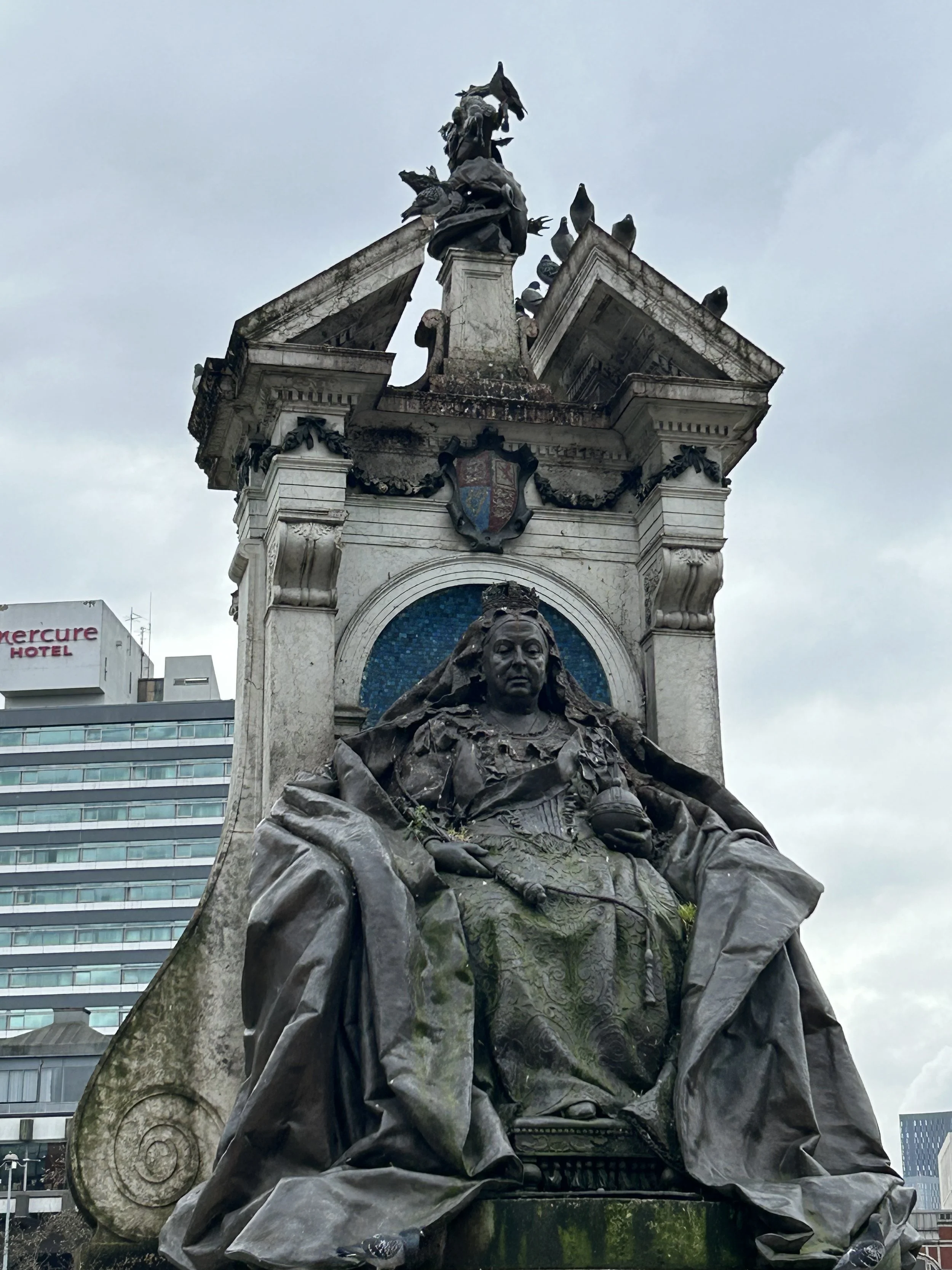 Statue of a seated woman wearing a crown, surrounded by decorative elements, with a coat of arms above, atop a stone pedestal in an urban setting.