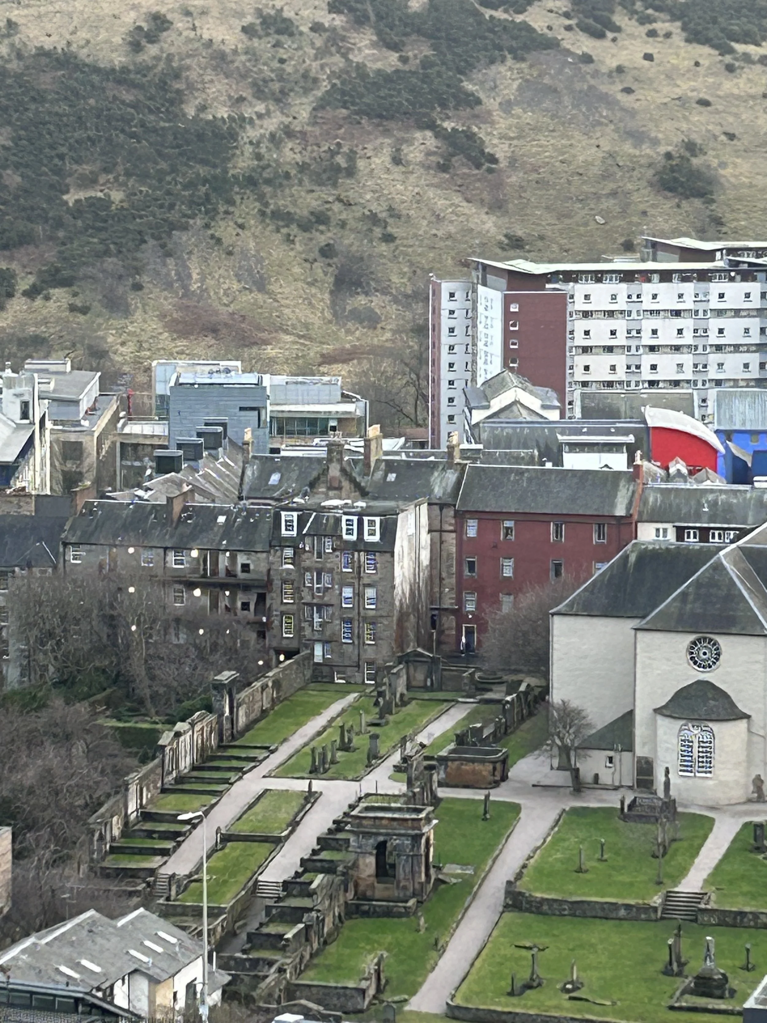 Aerial view of a cityscape with old and modern buildings, a church, and a green cemetery in the foreground with pathways and gravestones, hills in the background.
