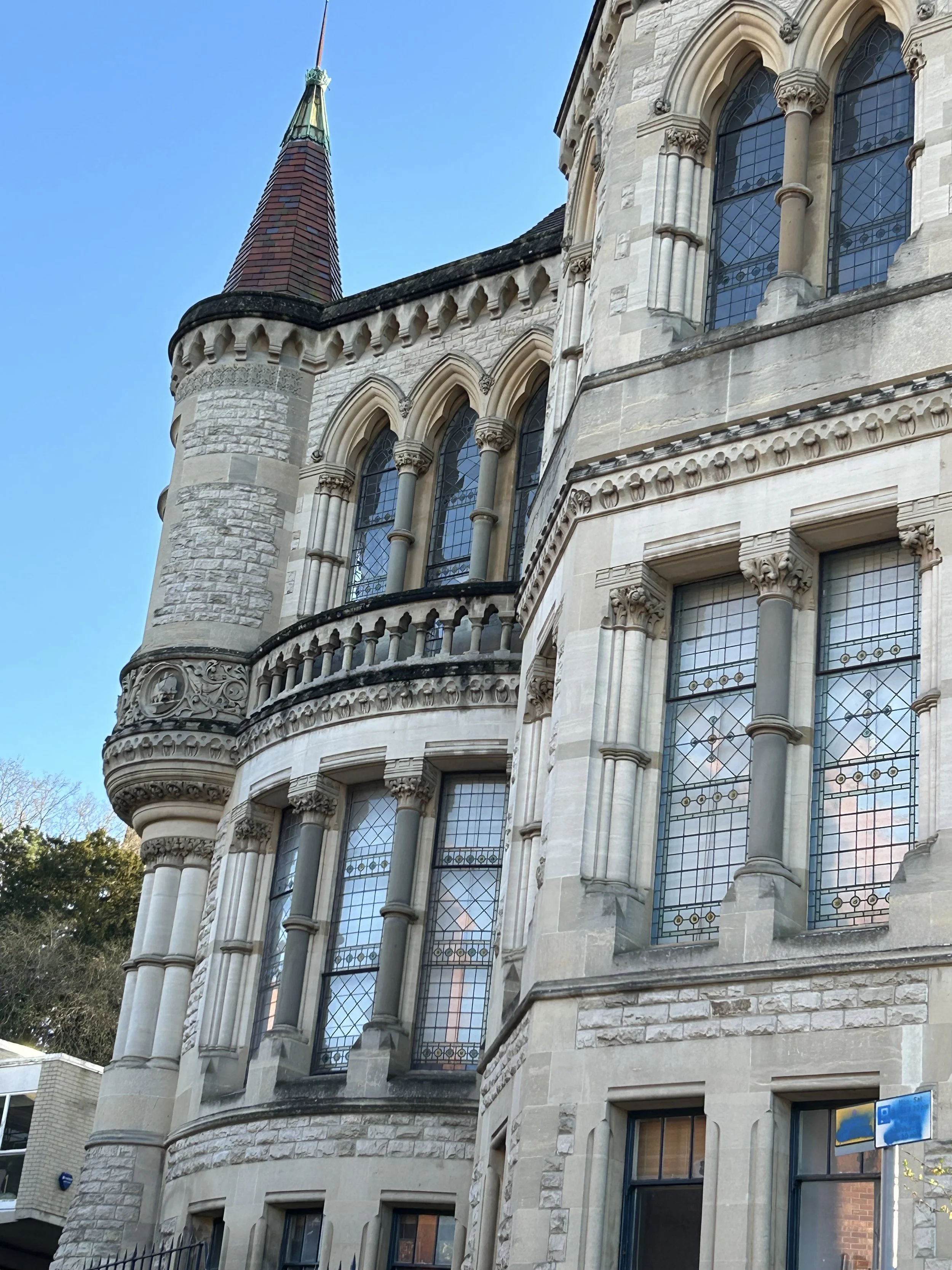 Close-up view of an ornate historic castle-style building with stone walls, arched windows with stained glass, and a sharp, pointed tower with a red and green roof against a clear blue sky.