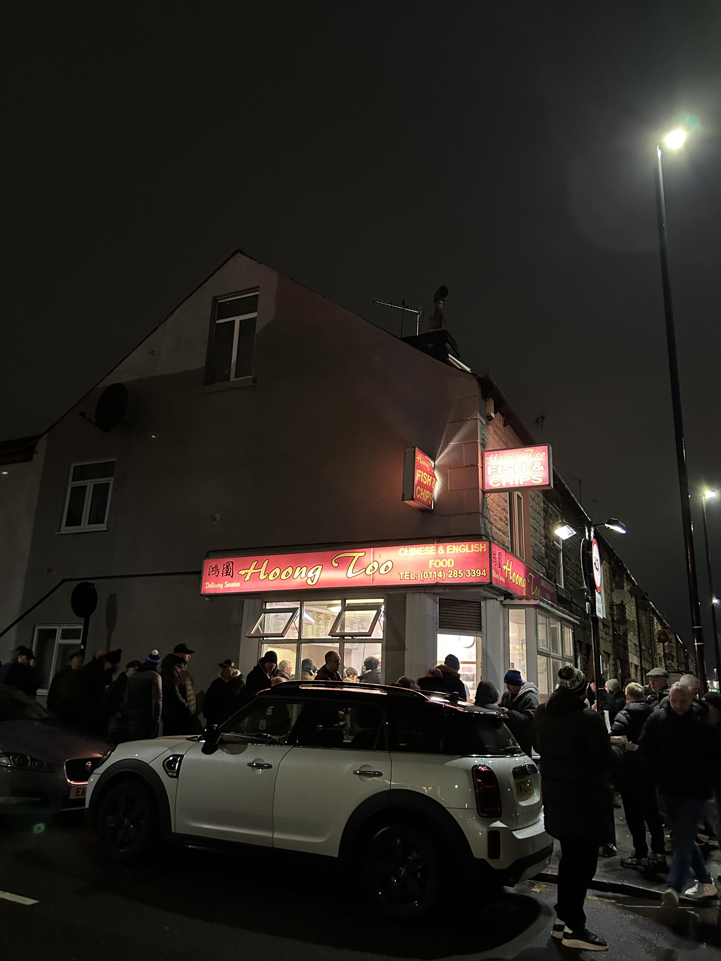 Night scene outside Hoong Too restaurant with a crowd of people waiting and walking on the sidewalk. The restaurant has red neon signs with English and Chinese characters, and a white car parked in front. The building is a two-story structure, and th