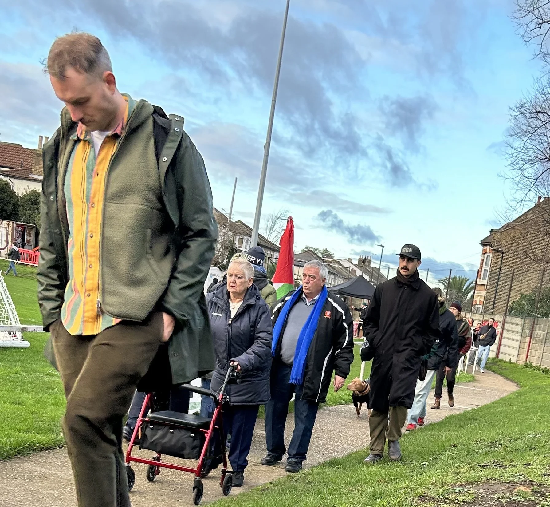 People walking in a line outdoors on a cloudy day, some with dogs, in a park or neighborhood with houses and a flag in the background.