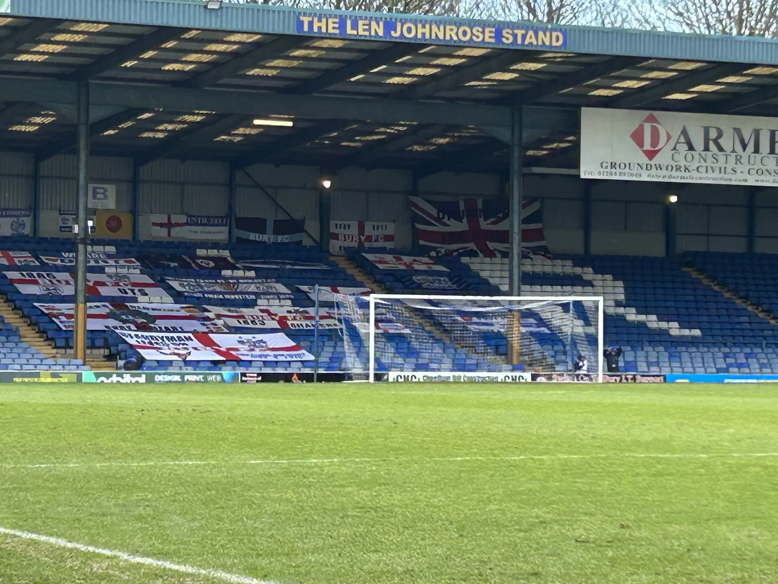 A soccer stadium with a goalpost, empty seats, and banners, including one for the Len Johnrose stand and England flags, under a blue roof.