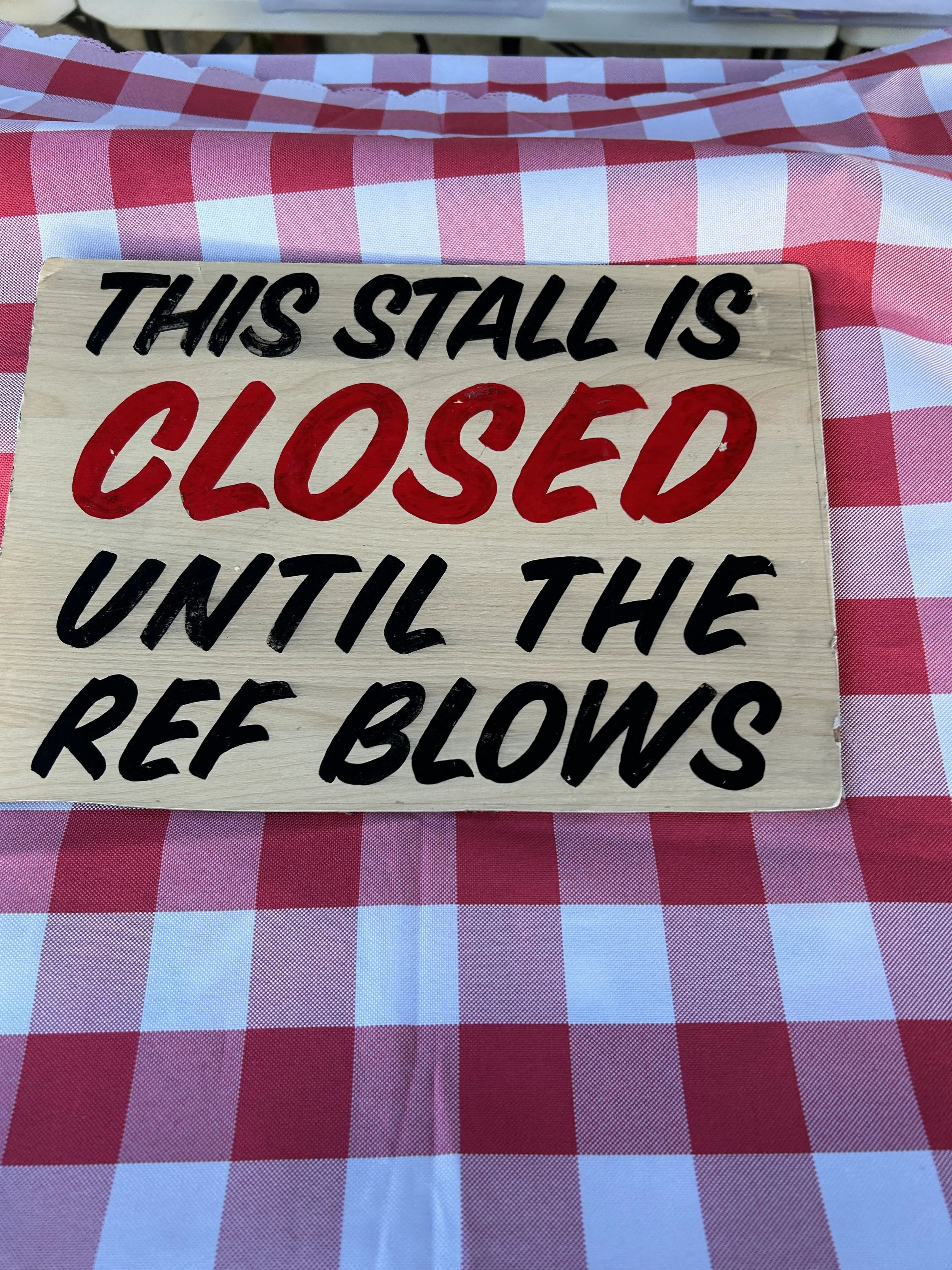 A wooden sign with black and red lettering reading 'This stall is closed until the ref blows,' placed on a red and white checkered tablecloth.