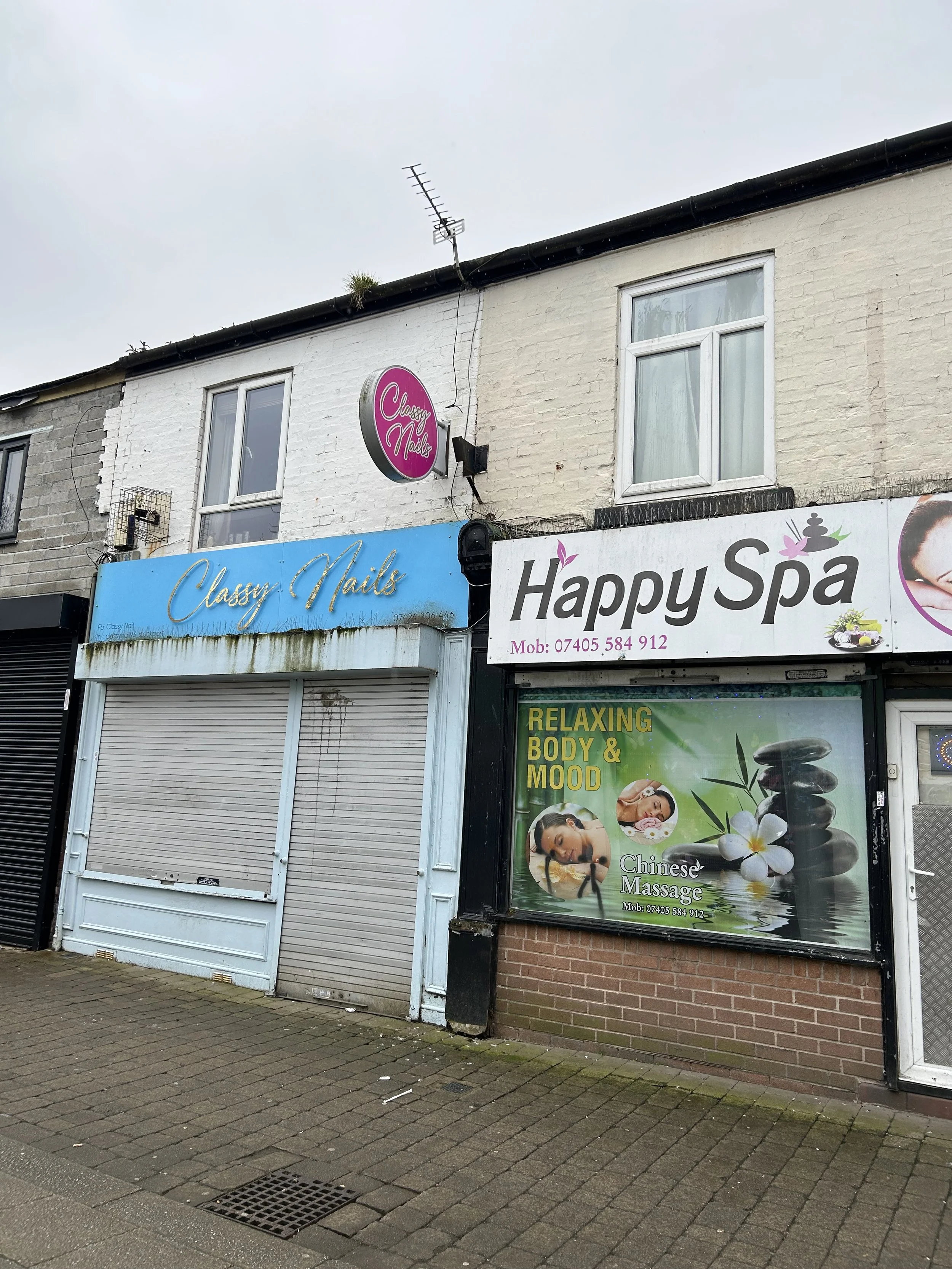 A storefront with signs for 'Classy Nails' and 'Happy Spa,' featuring massage and relaxation services advertised as Chinese massage with images of spa stones, flowers, and people relaxing.