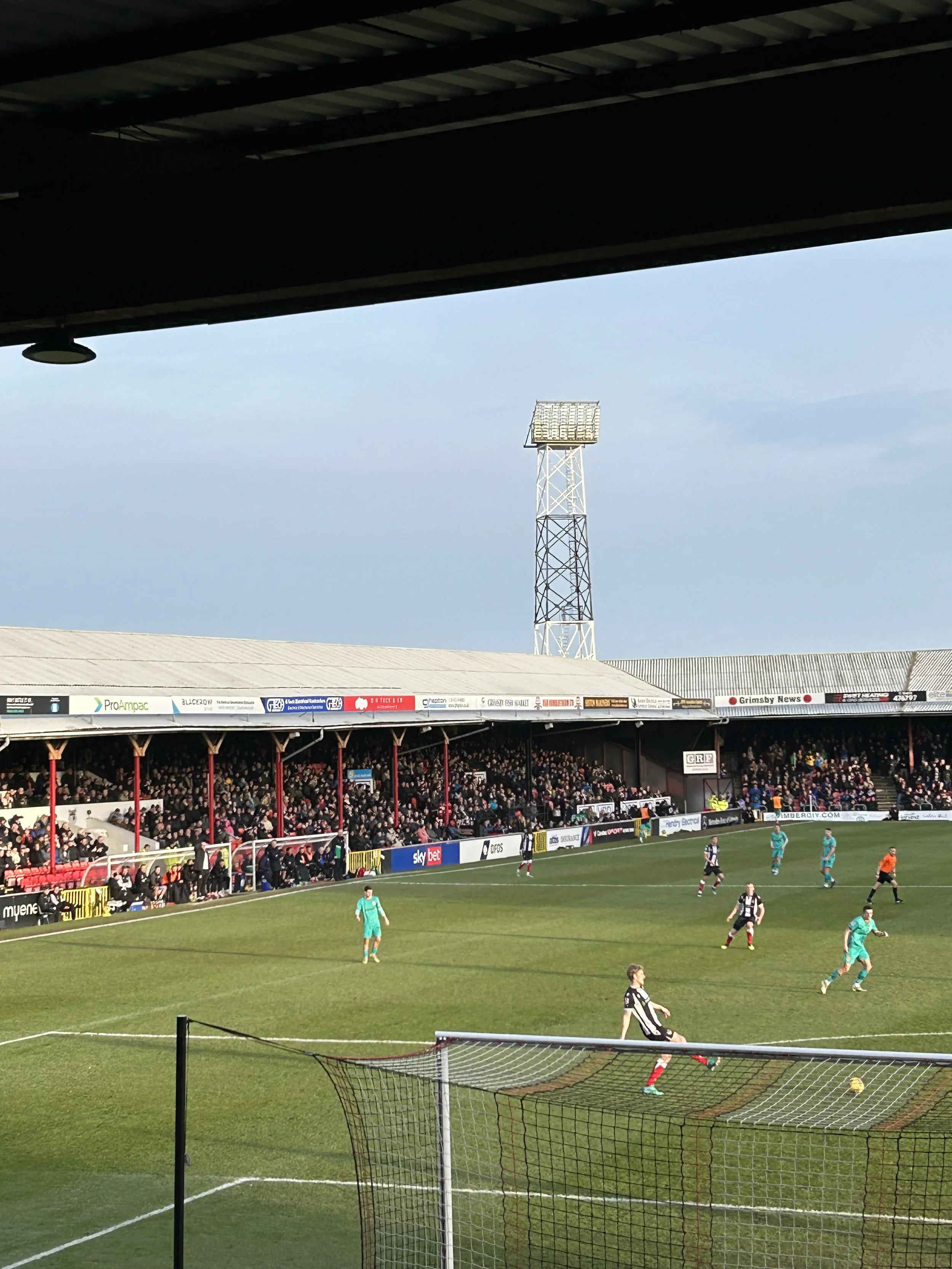 A soccer match taking place in a stadium with spectators in the stands. Players are on the field, some in black and white striped jerseys and others in teal uniforms. The goal and net are visible in the foreground, and a tall lighting tower is seen i