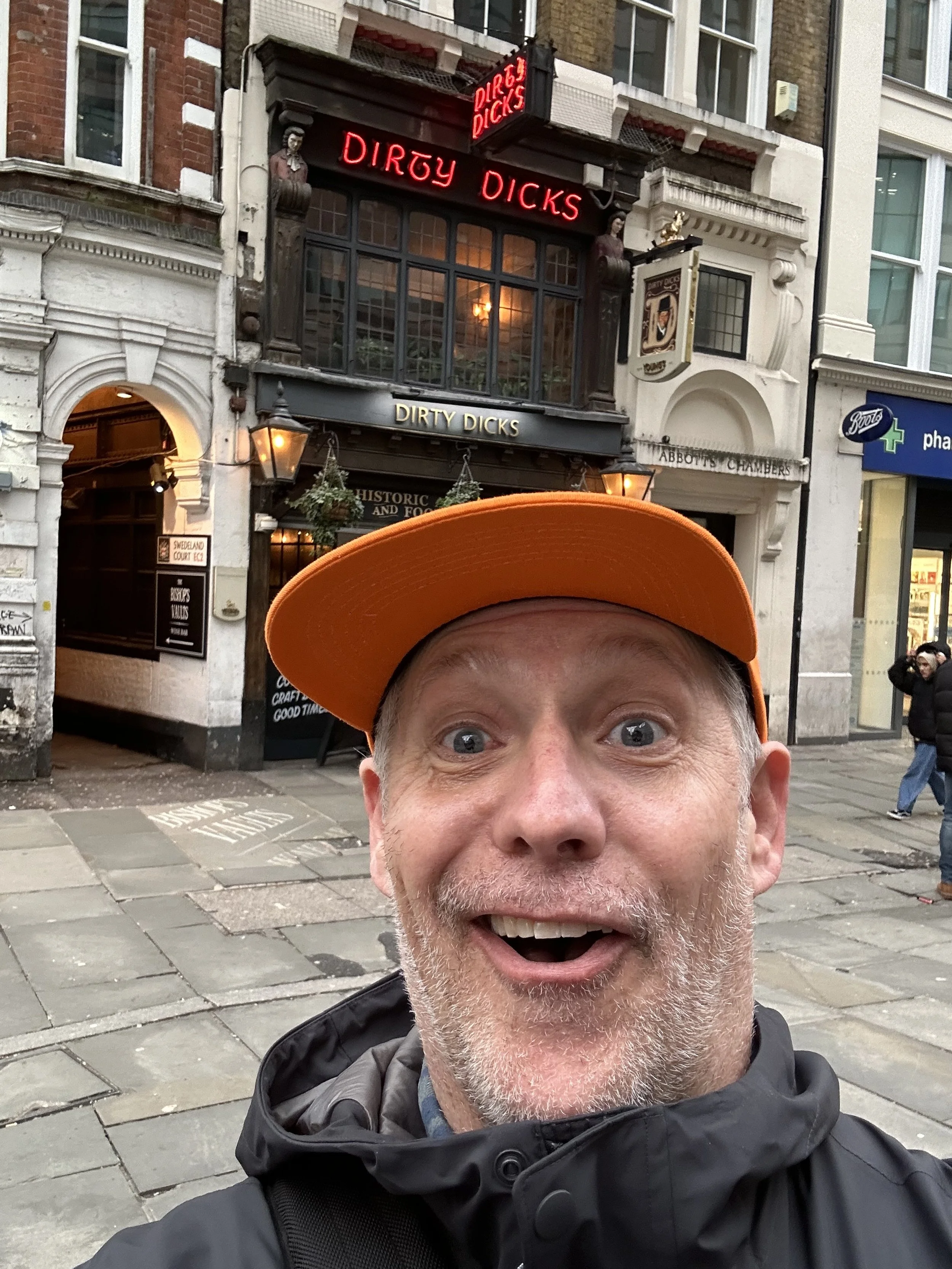 Author, Todd Smith with a surprised expression wearing an orange cap and black jacket taking a selfie in front of the Dirty Dicks pub on a city street.