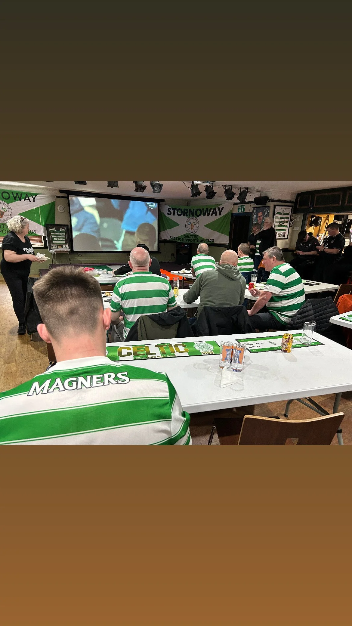 Group of people watching a soccer game on a large screen in a decorated club with Celtic-themed banners and table settings.