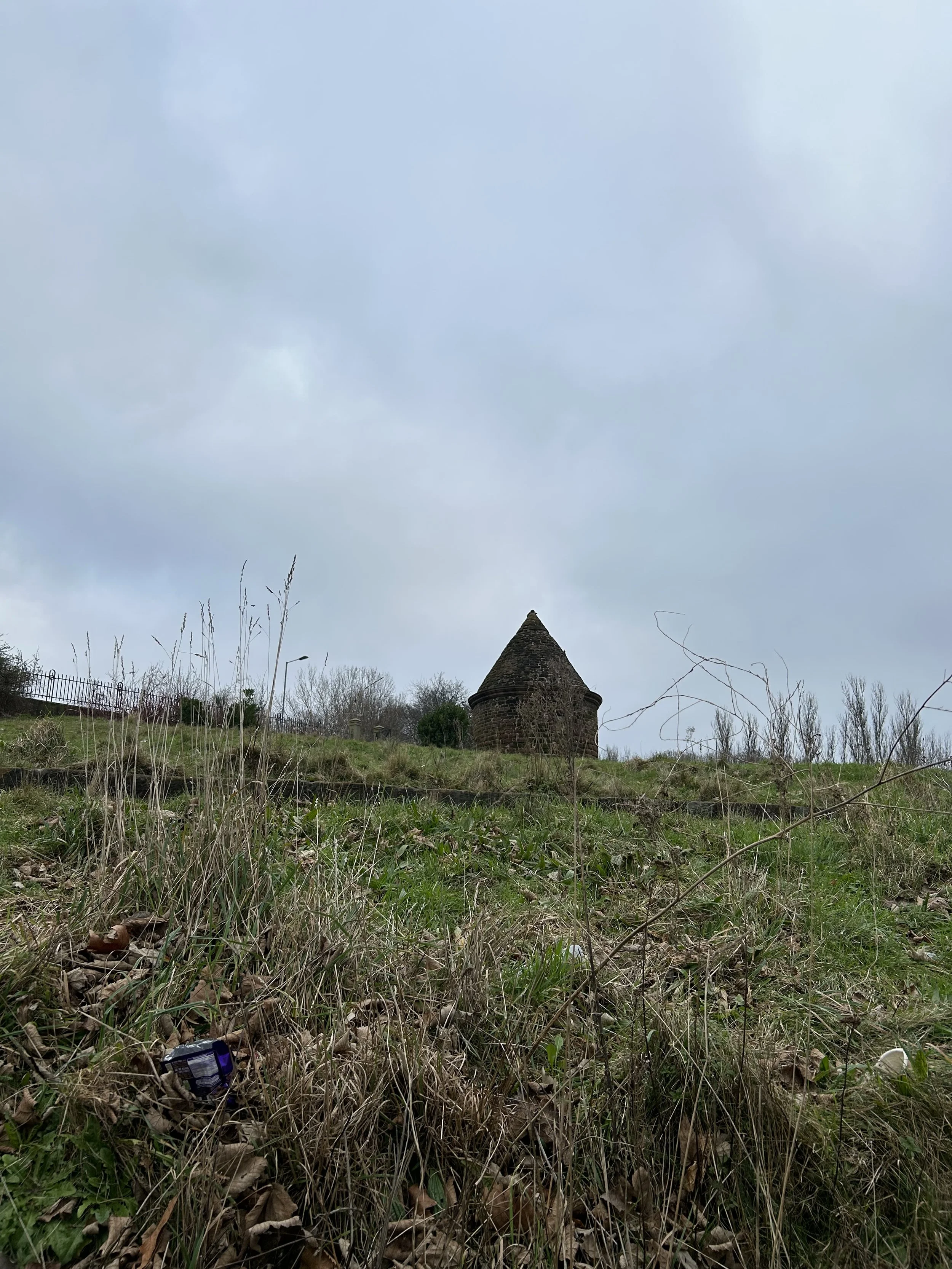 An old stone tower with a conical roof atop a grassy hill, with overcast skies and sparse trees in the background, and some scattered trash in the foreground.