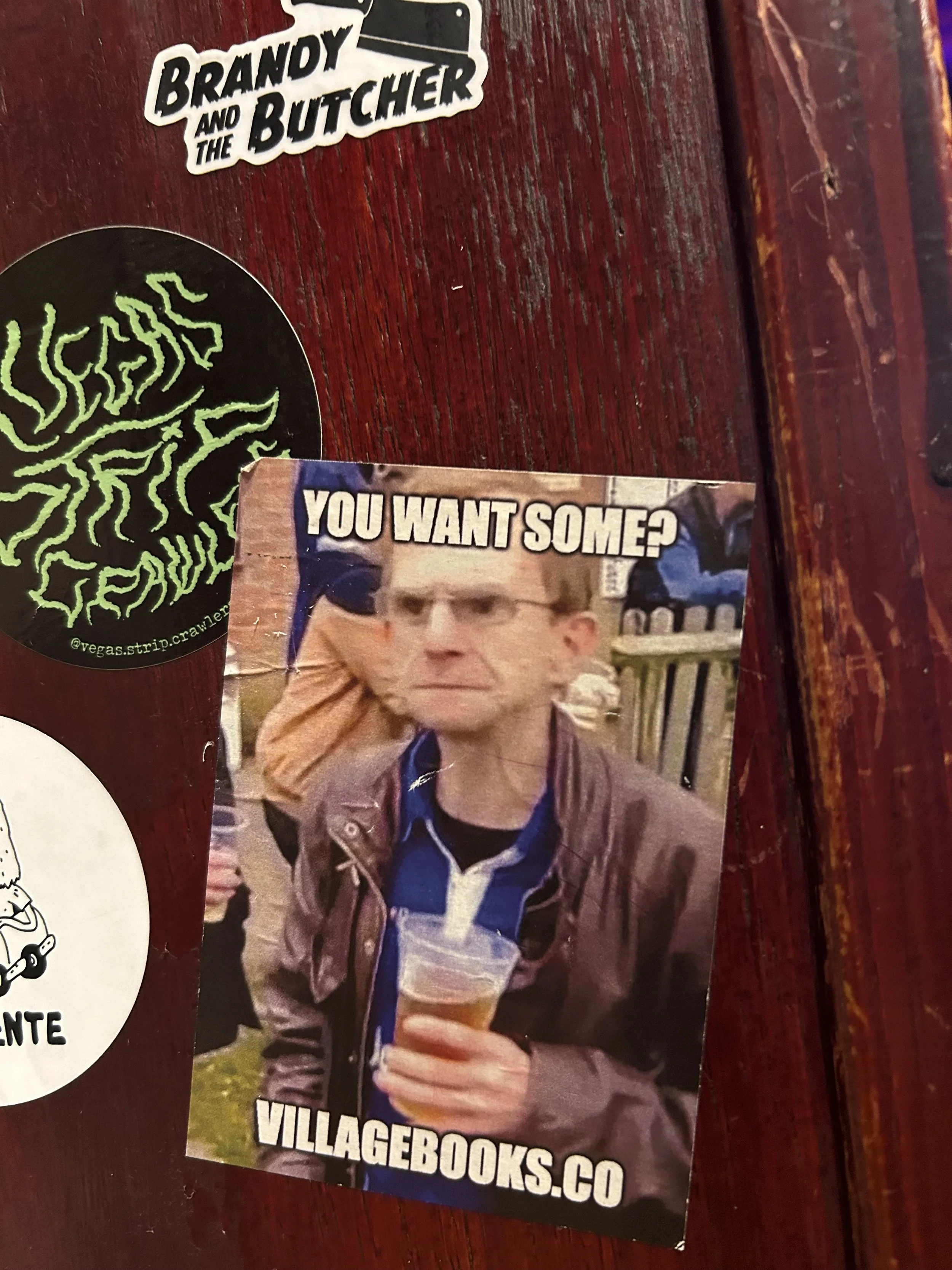 A sticker with a photo of a man holding a plastic cup of beer, looking serious, and the text 'You Want Some?' above him, and 'VILLAGEBOOKS.CO' below.