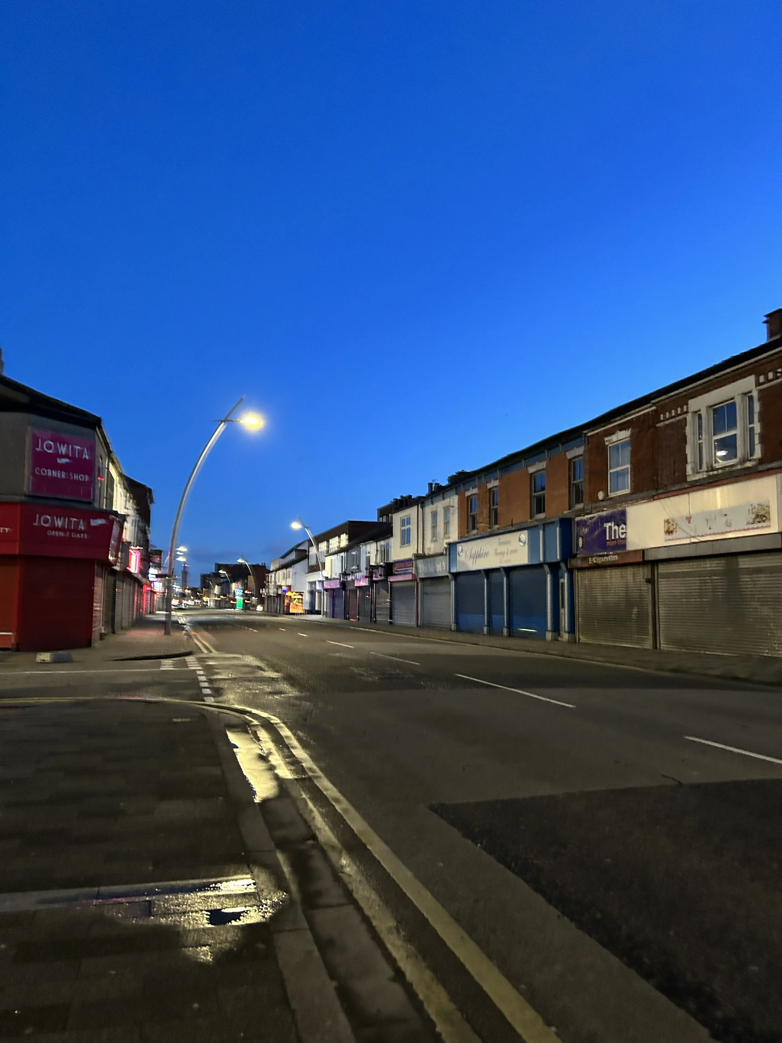 Empty street with closed shops, streetlights on, during dusk or dawn, with a clear blue sky.