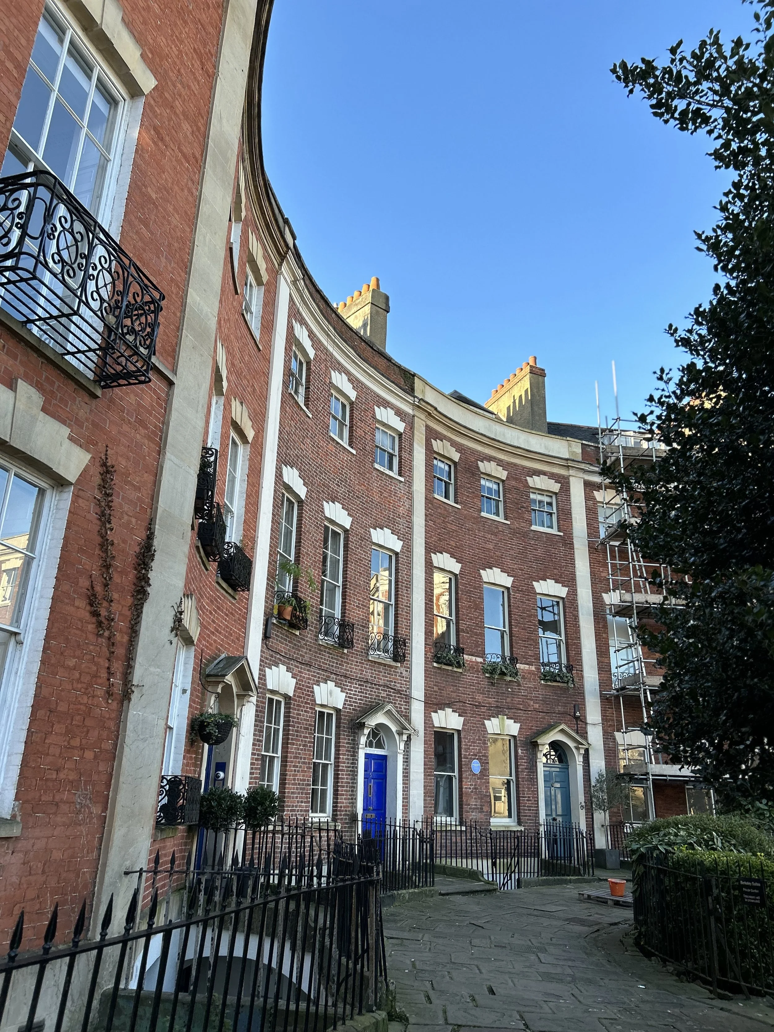 A curved brick residential building with multiple floors, white window frames, small balconies with black iron railings, potted plants, and a blue door, under a clear blue sky.