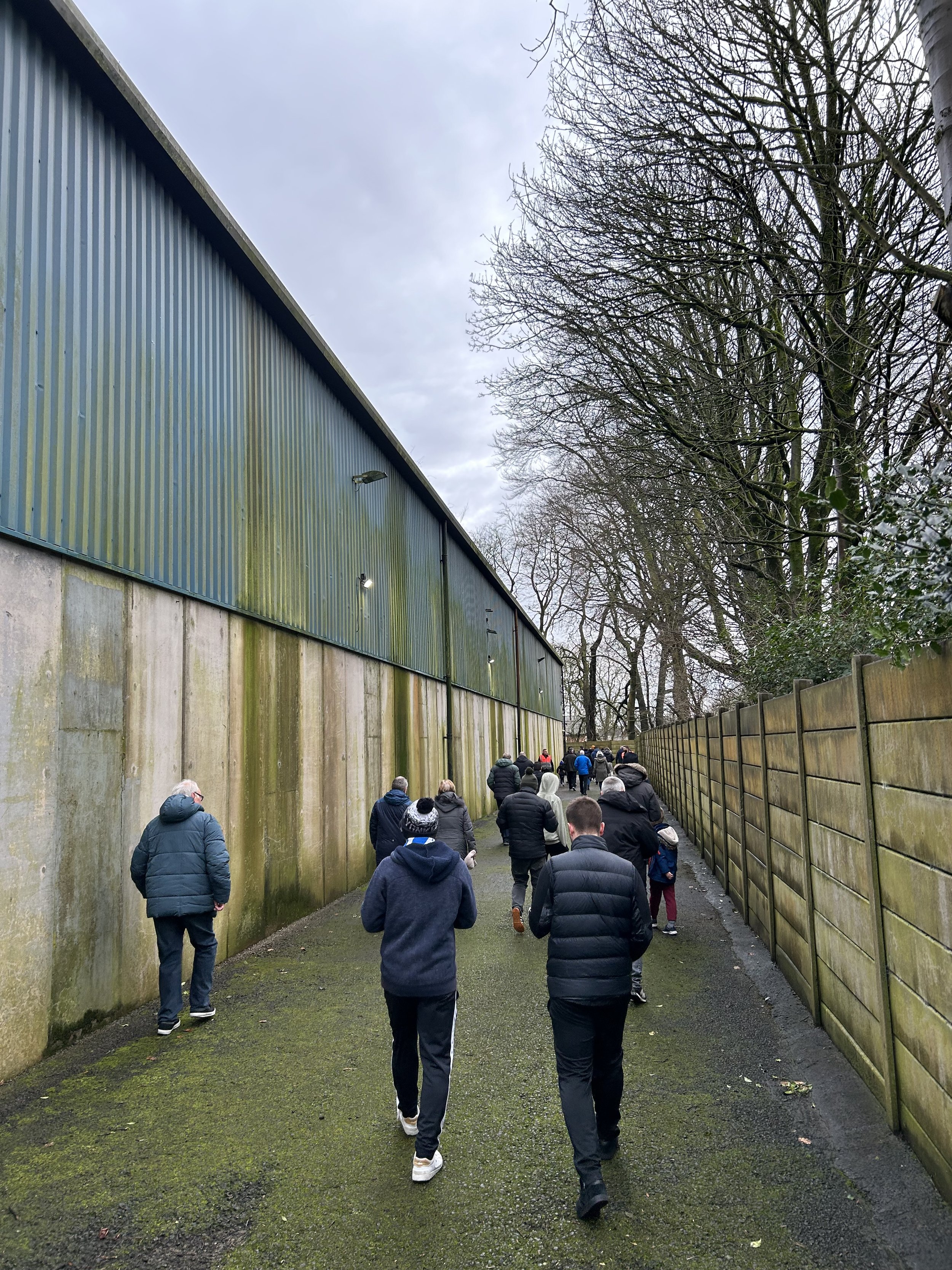 Group of people walking along a narrow outdoor pathway between a tall mossy concrete wall and a fence, with leafless trees overhead on a cloudy day.