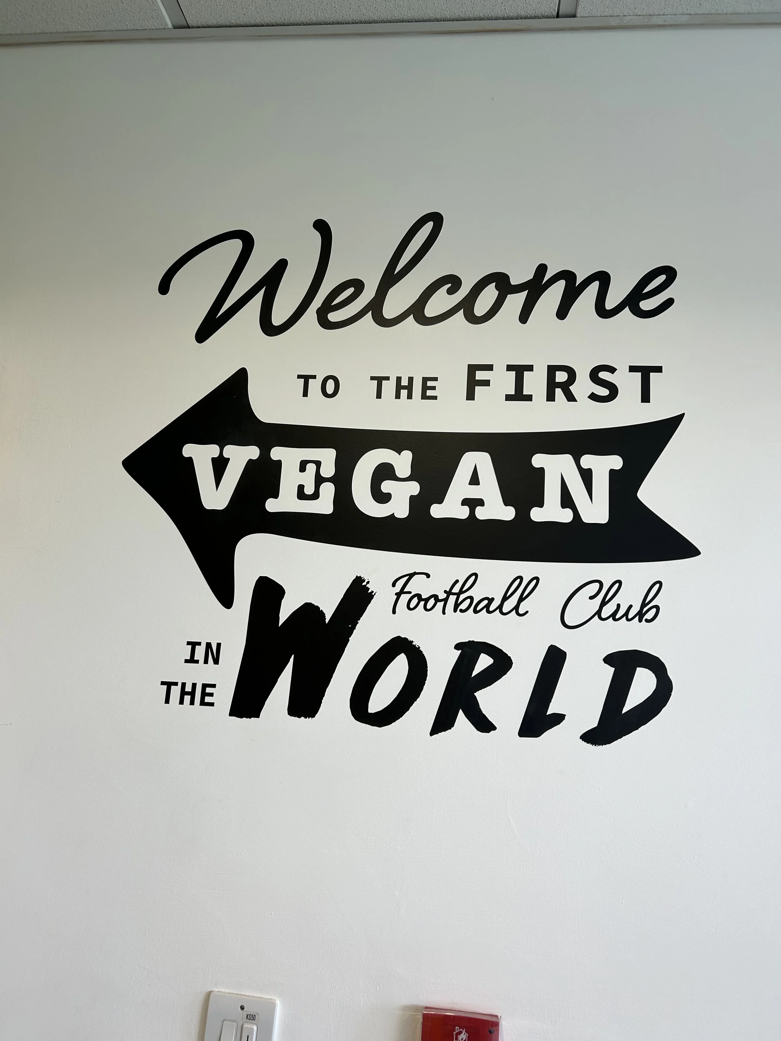 Wall art with the text "Welcome to the First Vegan Football Club in the World" in black and white lettering, with a large black arrow behind the word "Vegan."