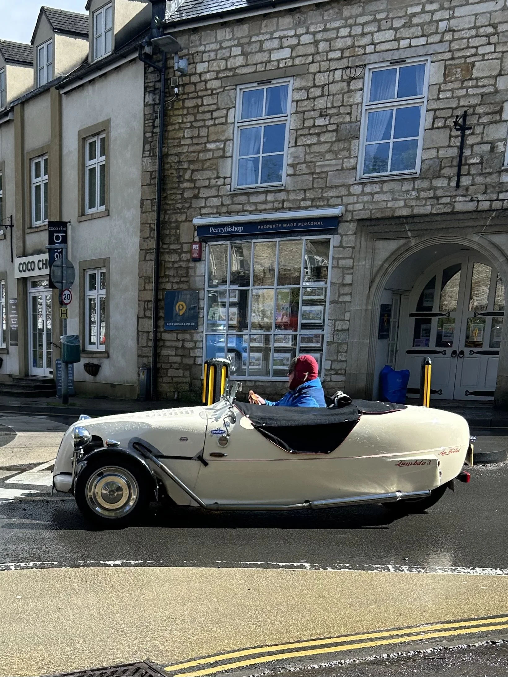 A vintage cream-colored three-wheeled car with a black sunshade parked on the street. A person wearing a red cap and blue jacket is sitting inside, looking at their phone. There are buildings with storefronts behind the car, including a property agen
