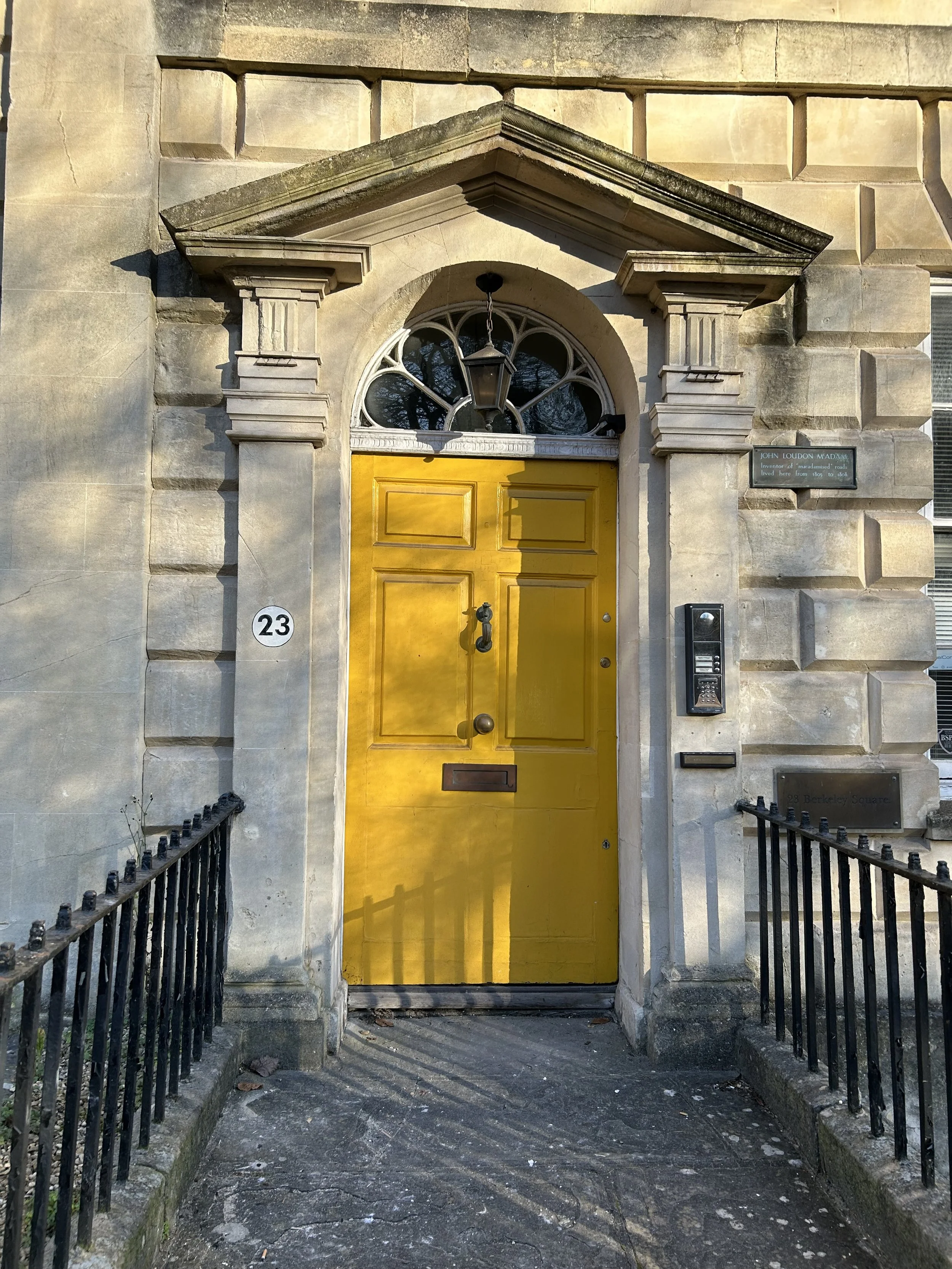 Front door with bright yellow paint, arched window above, hanging lantern light, black intercom and mail slot on the right, iron railings on sides, stone steps and walls, house number 23, and a green plaque.