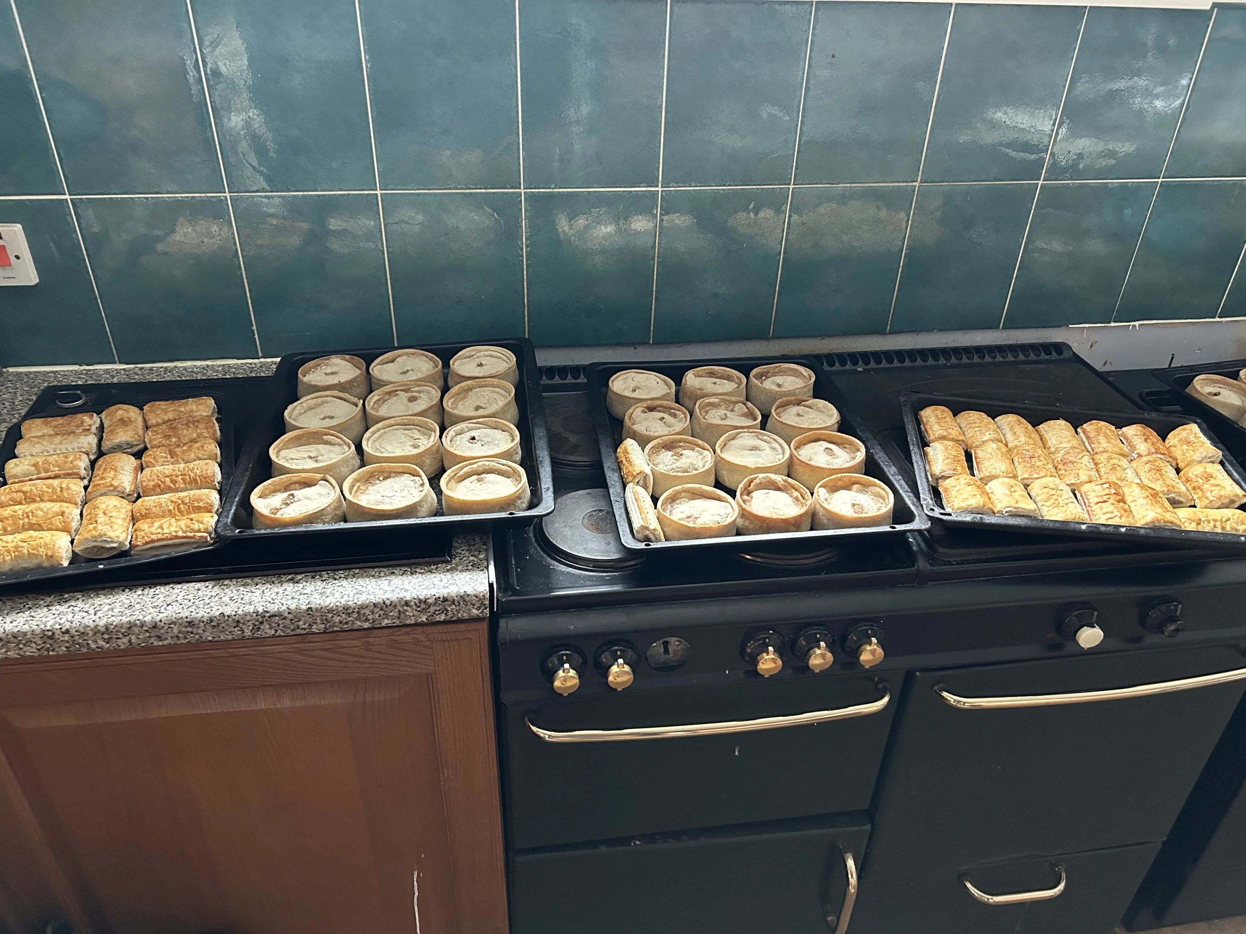 Ovens filled with trays of baked pastry rolls and small pie-like desserts cooling on a black stovetop.