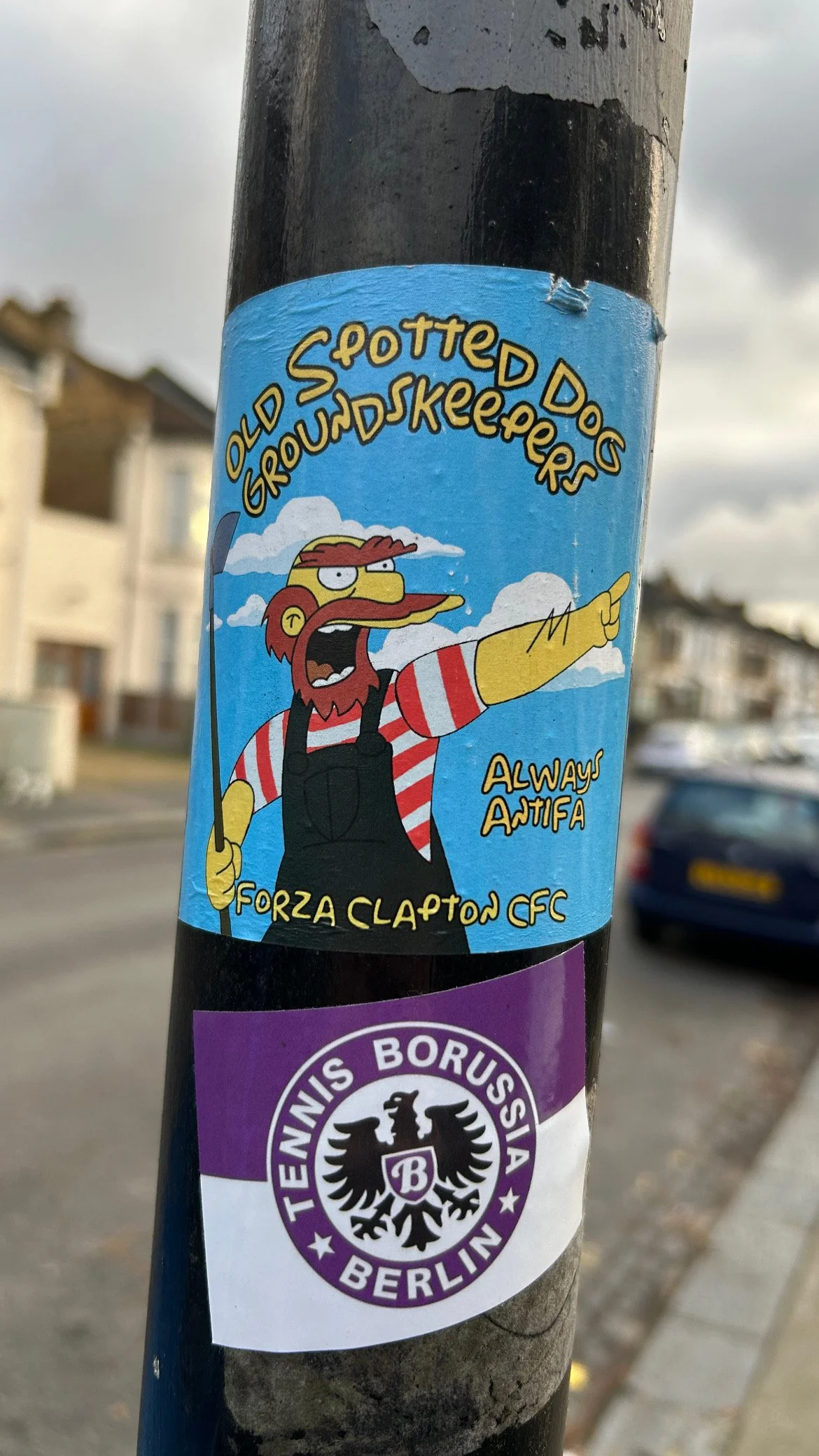 Sticker of Groundskeeper Willie from The Simpsons, wearing a red and white striped shirt and black overalls, pointing and shouting, with the text "Old Spotted Dog Groundskeepers Always AntiFa Forza Clapton CFC." Below is a purple and white sticker wi
