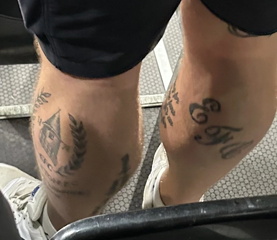 Close-up of a person's tattooed legs, wearing black shorts and white sneakers, standing on a rubber gym floor.