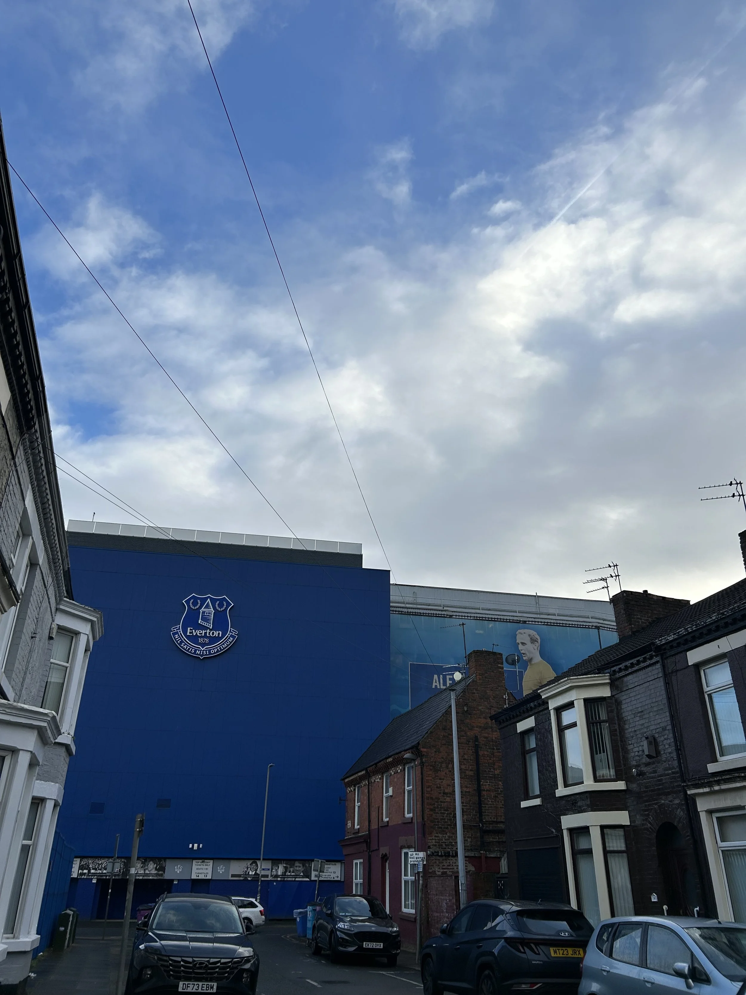 Street view showing residential houses and a large blue building with the Everton football club logo and a billboard of a player, under a partly cloudy sky.