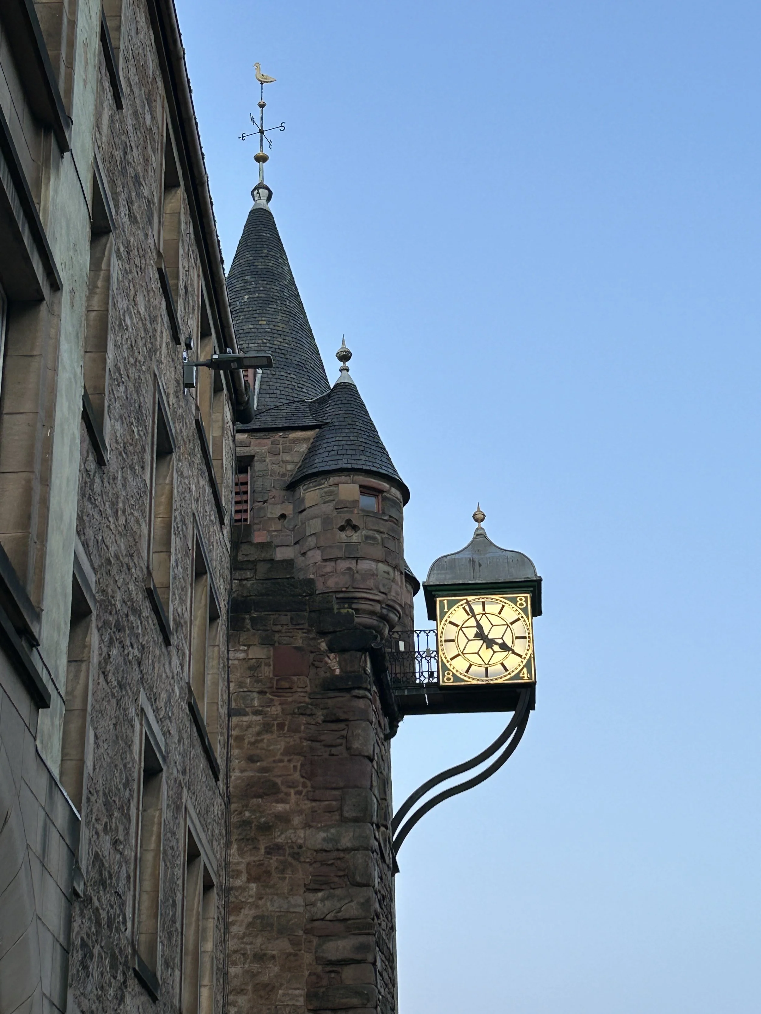 A historic stone building with a clock tower, featuring pointed turrets and a weather vane on top, against a clear blue sky.