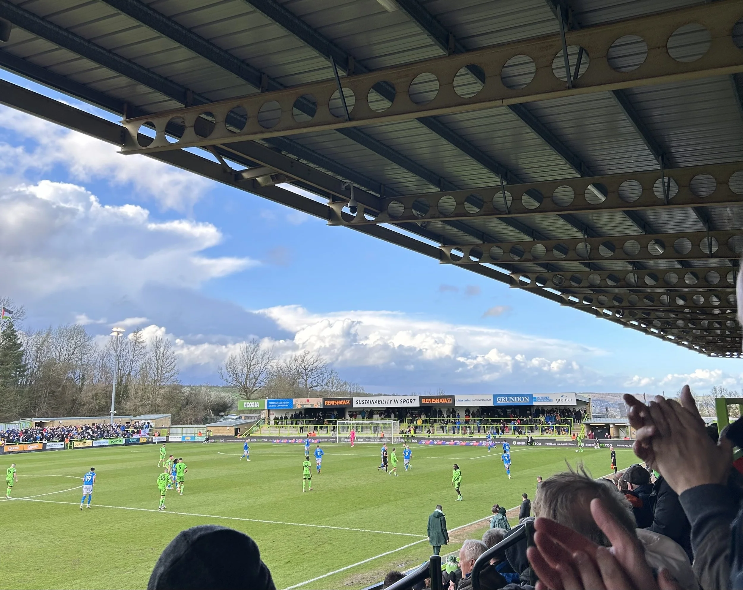 A soccer match in a stadium with players in green and blue uniforms on the field, spectators in the stands, and a partly cloudy sky overhead.