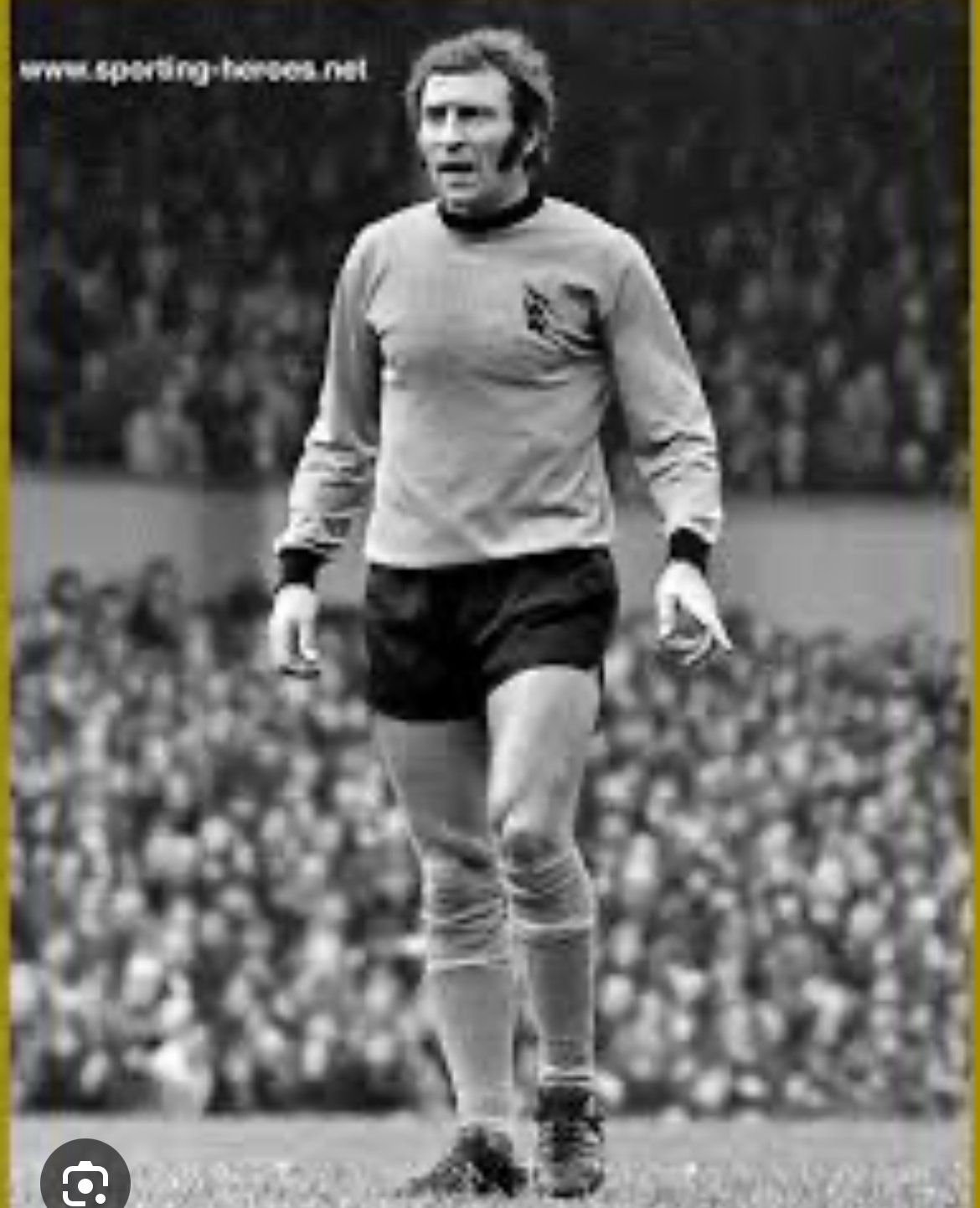Black and white photo of a soccer player on the field, wearing a long-sleeve jersey, shorts, and socks, with a crowd in the background.