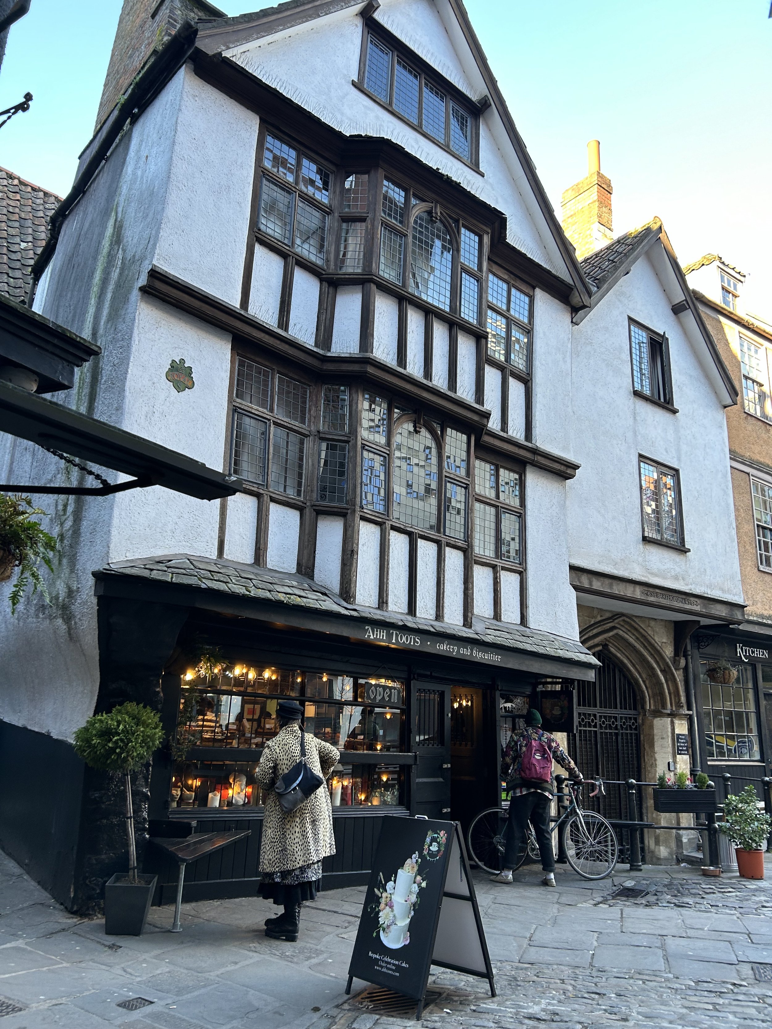 A historic three-story building with Tudor-style architecture featuring dark wooden framing and white stucco walls. The ground floor is a bakery and cafe named 'Ahh Toots' with large windows and outdoor seating. Two people are standing outside, one w