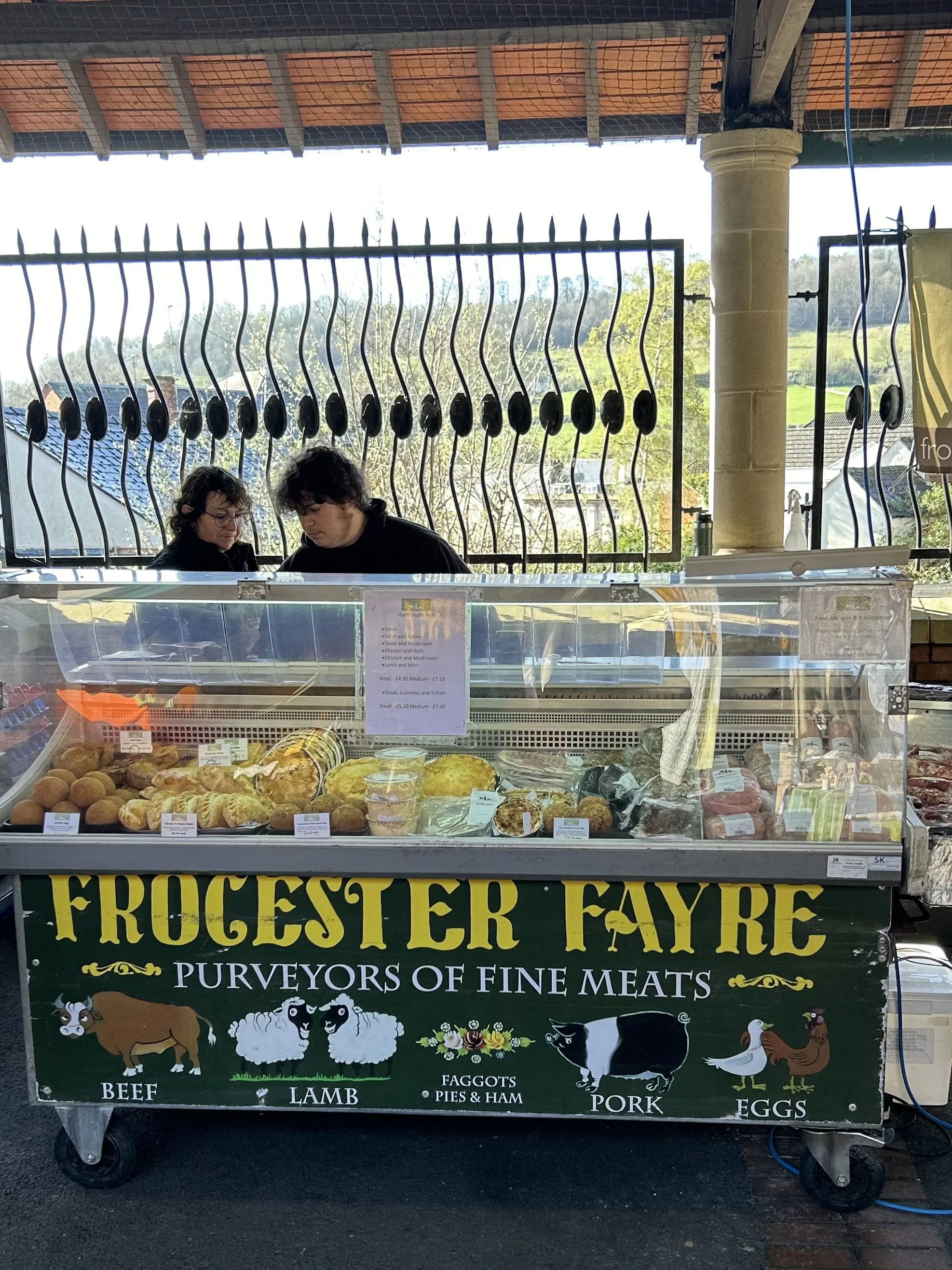 A market stand with a sign reading "Froster Fayre Purveyors of Fine Meats" featuring illustrations of beef, lamb, pigs, pork, eggs, and faggots, with various meats and baked goods displayed inside a glass case.