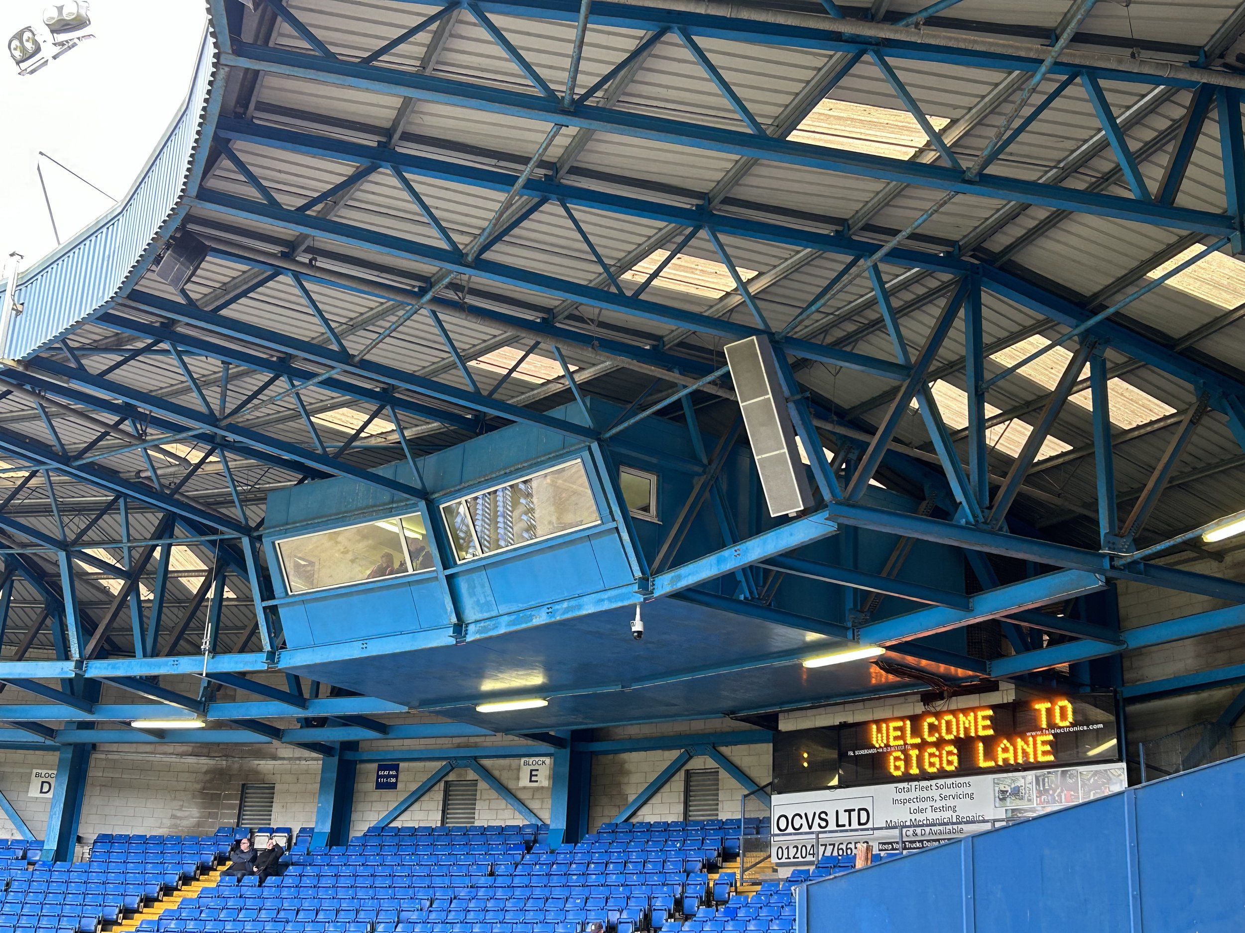 A section of a sports stadium with blue seats, a metal roof, and a large digital sign that reads 'WELCOME TO GIGG LANE'.