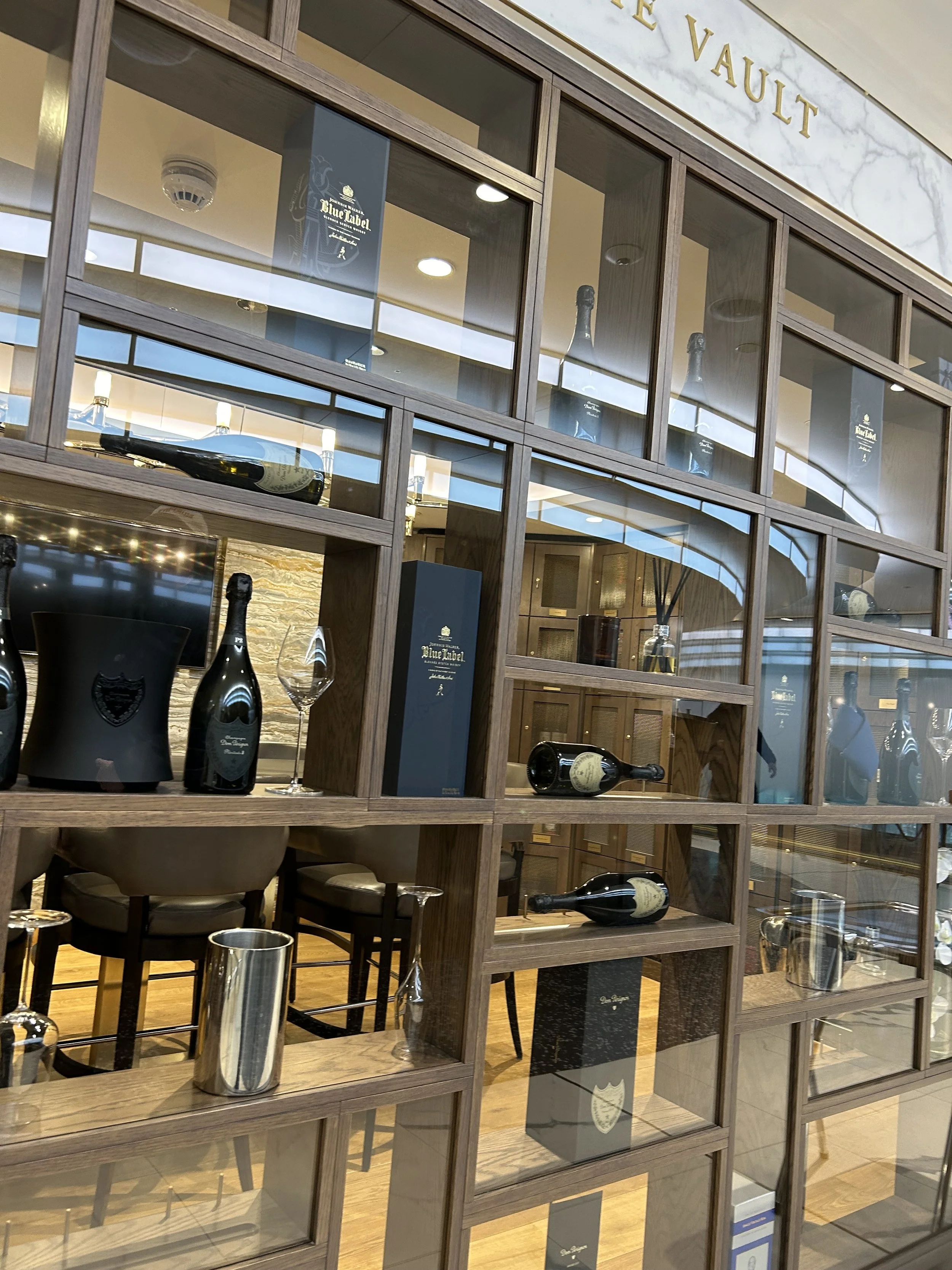 Display of assorted bottles of alcohol, including champagne and whiskey, arranged on wooden shelves behind glass at a bar or lounge area.