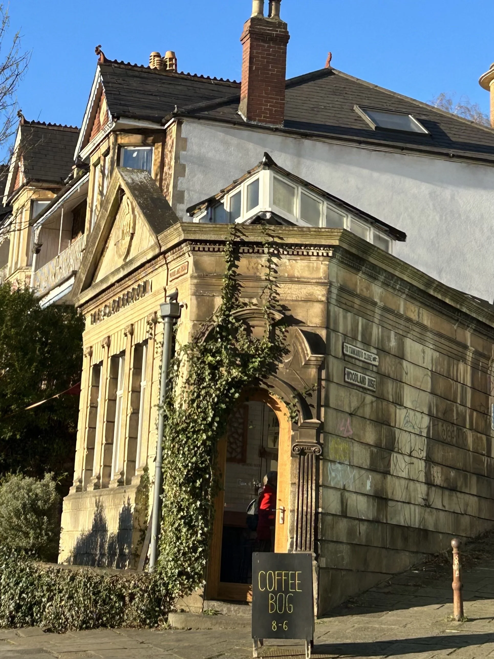 Old stone building with an arched entrance covered in ivy, a sign outside reads 'COFFEE BOG 8-6', with a person in a red jacket visible inside.