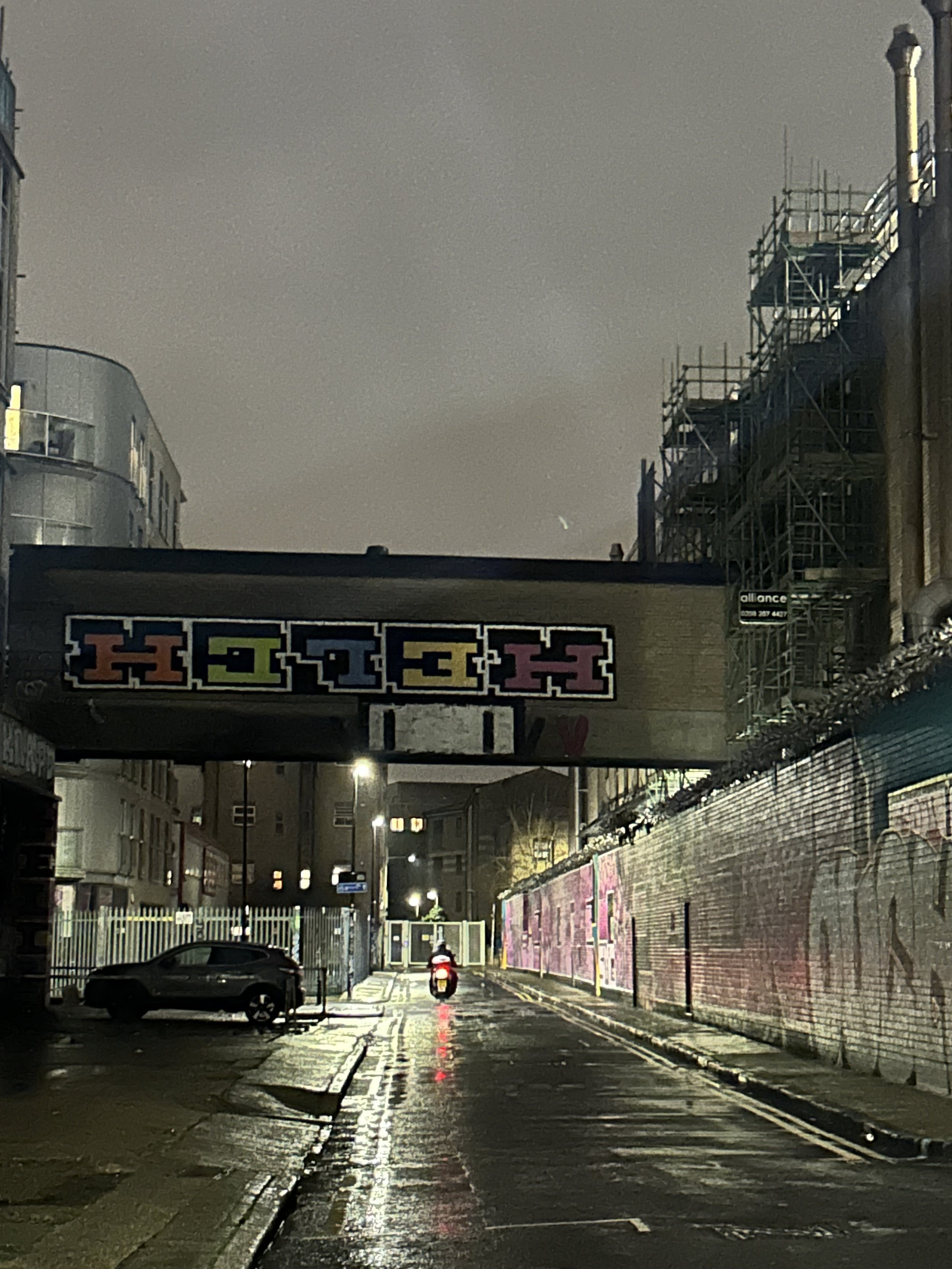 A wet city street at night with a single motorbike and a parked car. A colorful sign spells 'HOLA' on a bridge overhead, and there is graffiti on the right wall.