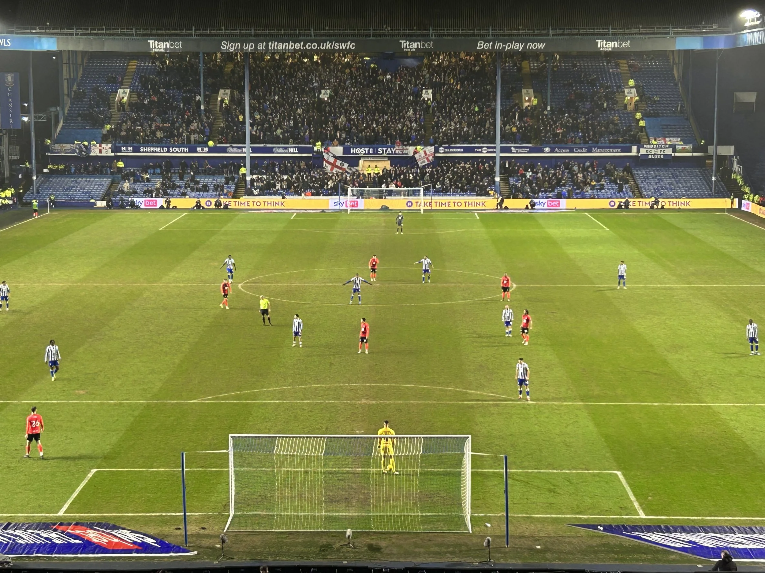 A soccer match in progress at a stadium with players on the field, spectators in the stands, and advertising banners around the perimeter.