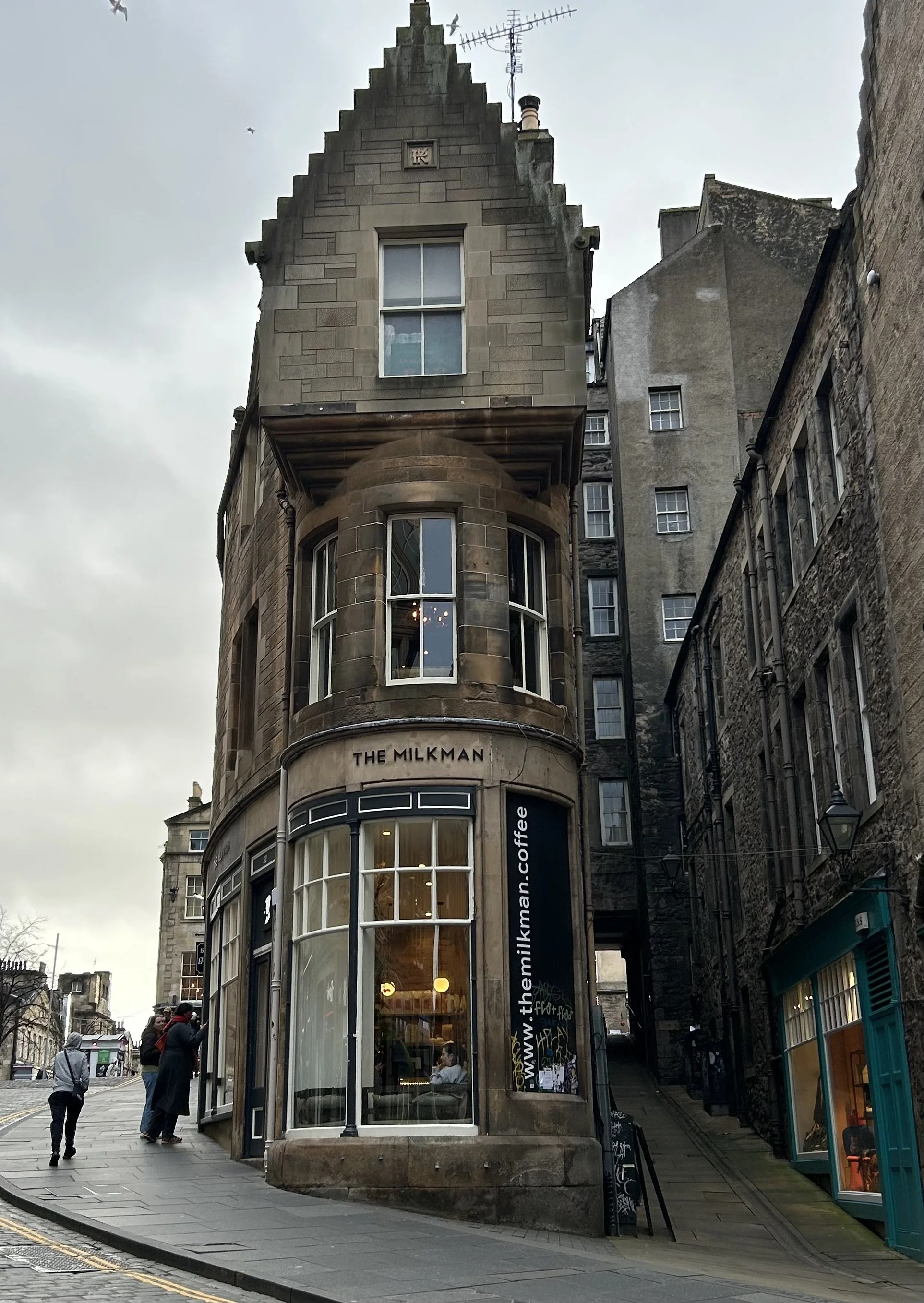 A multi-story building with a rounded corner at a street intersection. The ground floor is a coffee shop called 'The Milkman' with large windows, and inside customers are visible. The upper floors have windows, and the building has a distinctive gabl