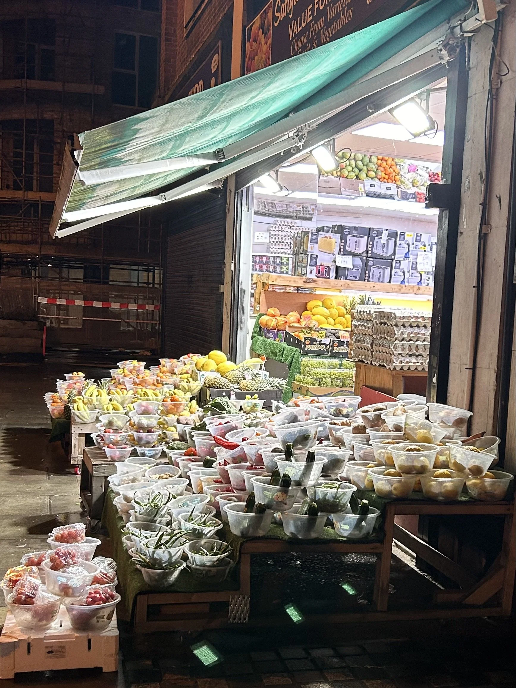 A fruit stand with various fresh fruits and vegetables on display outside a shop at night. The stand has plastic containers filled with fruit, and the shop behind has fruits like melons and oranges, along with boxes of eggs and kitchen appliances.