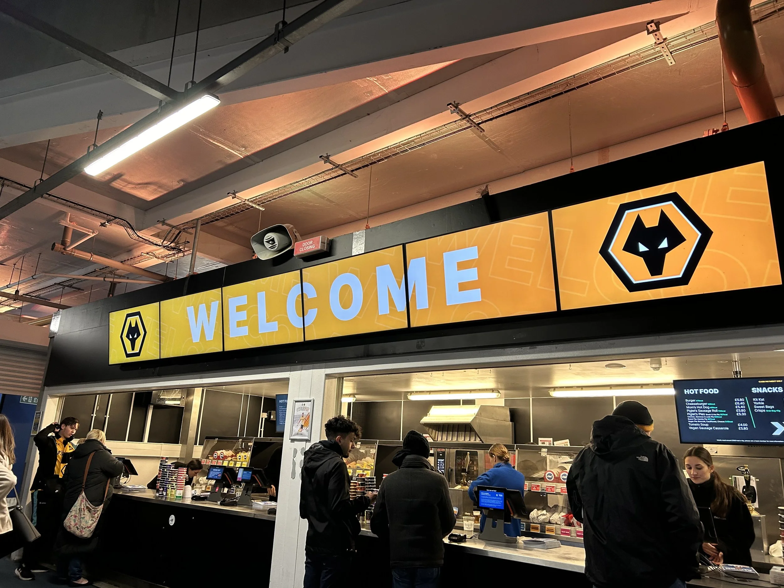 People ordering food at a counter under a large illuminated sign that reads 'WELCOME' with a wolf logo on either side, inside a fast food restaurant or cafeteria.