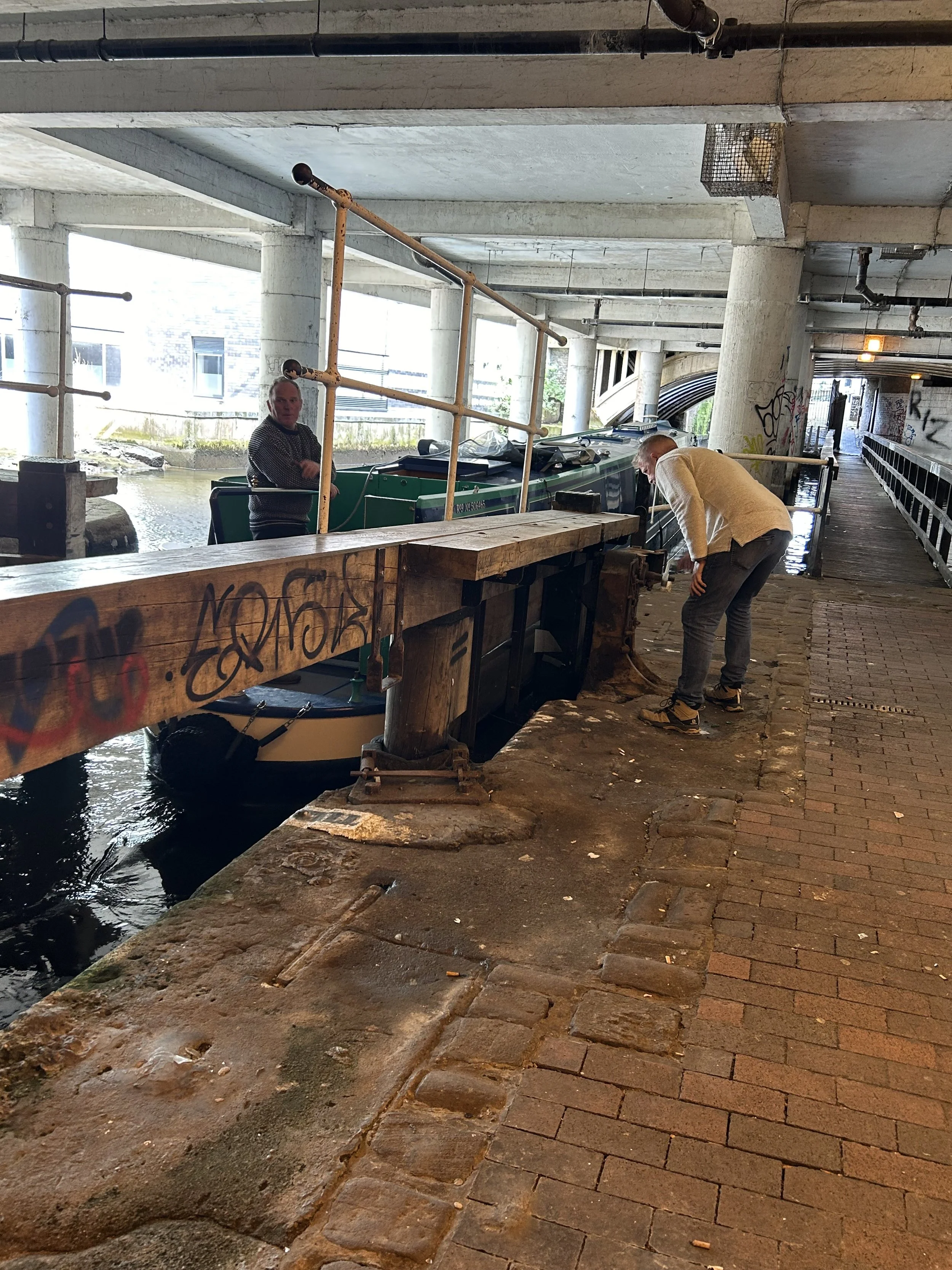 Two men are standing and bending over near a docked boat beneath a bridge. One man stands on the dock looking towards the camera while the other is inspecting the boat. The area is under a bridge with graffiti on the walls and a brick walkway.