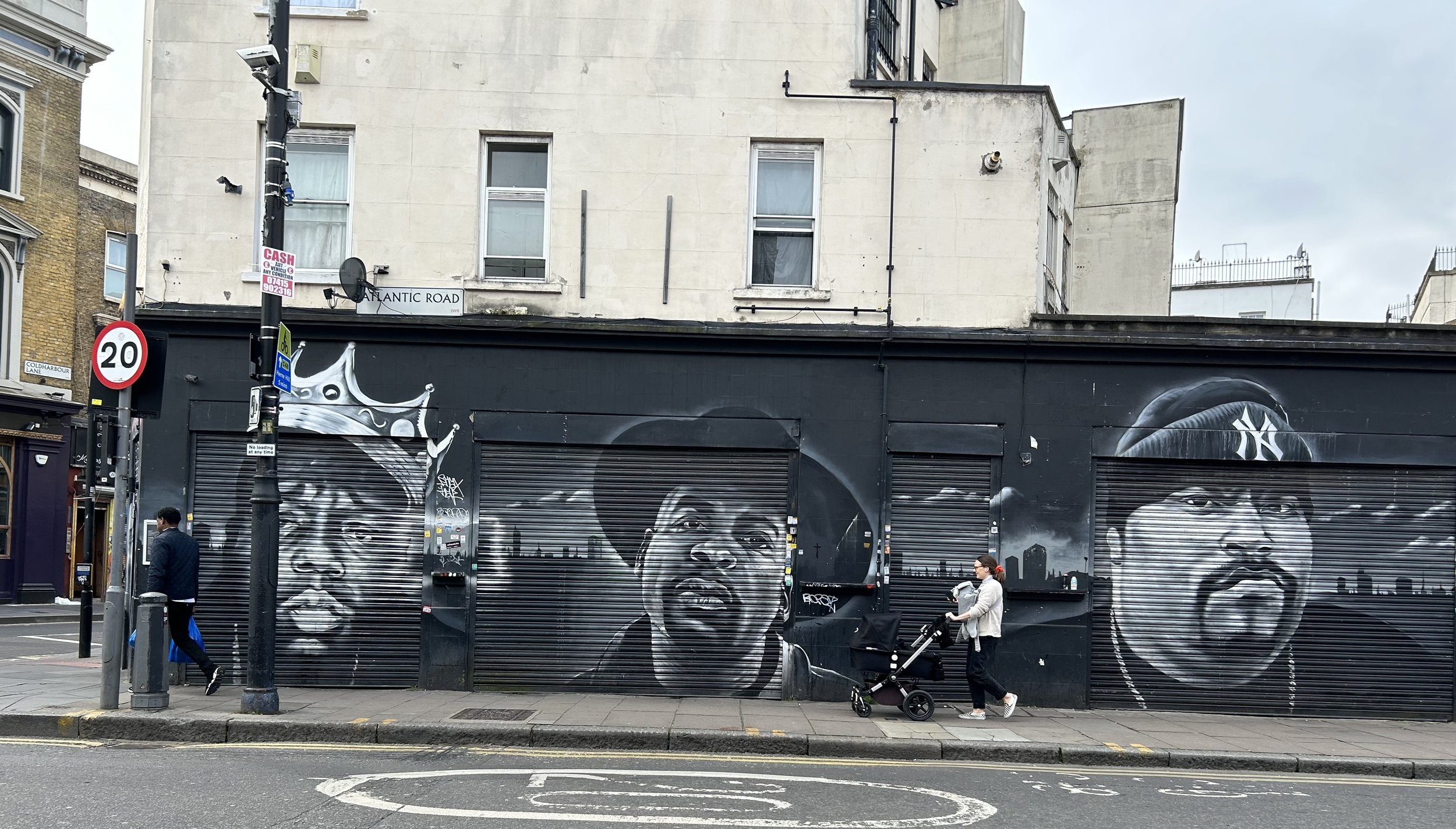 Street scene with mural graffiti of three black and white portraits of men on metal shutters, located on Atlantic Road. Pedestrians walking on sidewalk, one with a stroller, overcast sky.