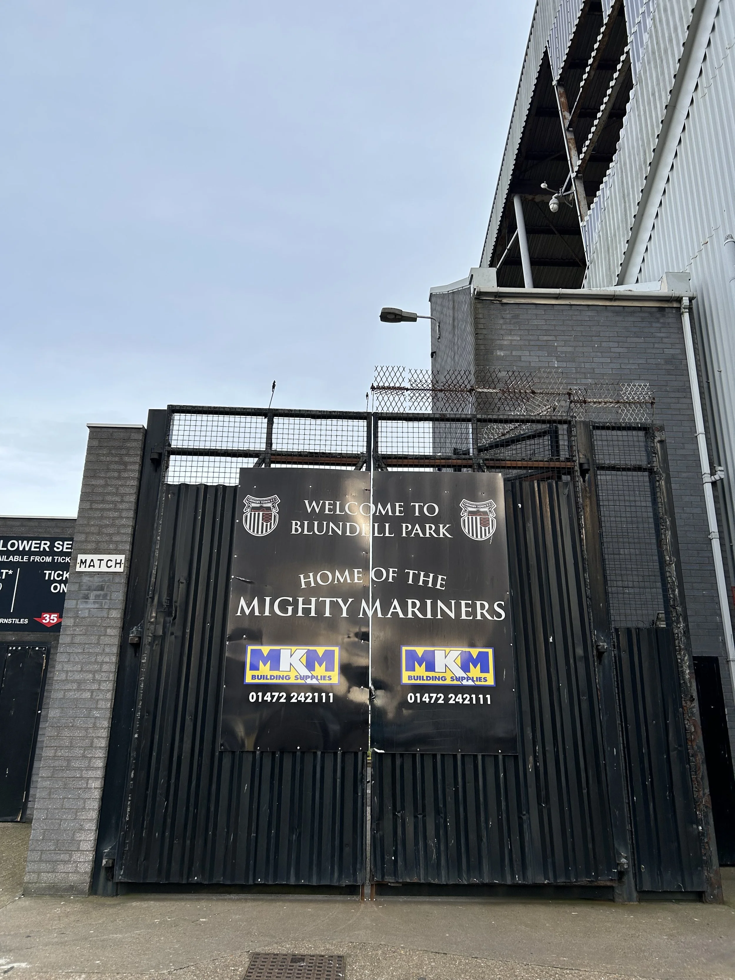 Black gate with a sign that reads "Welcome to Blundell Park, Home of the Mighty Mariners," with the MKM Building Supplies logo and contact number at the bottom.