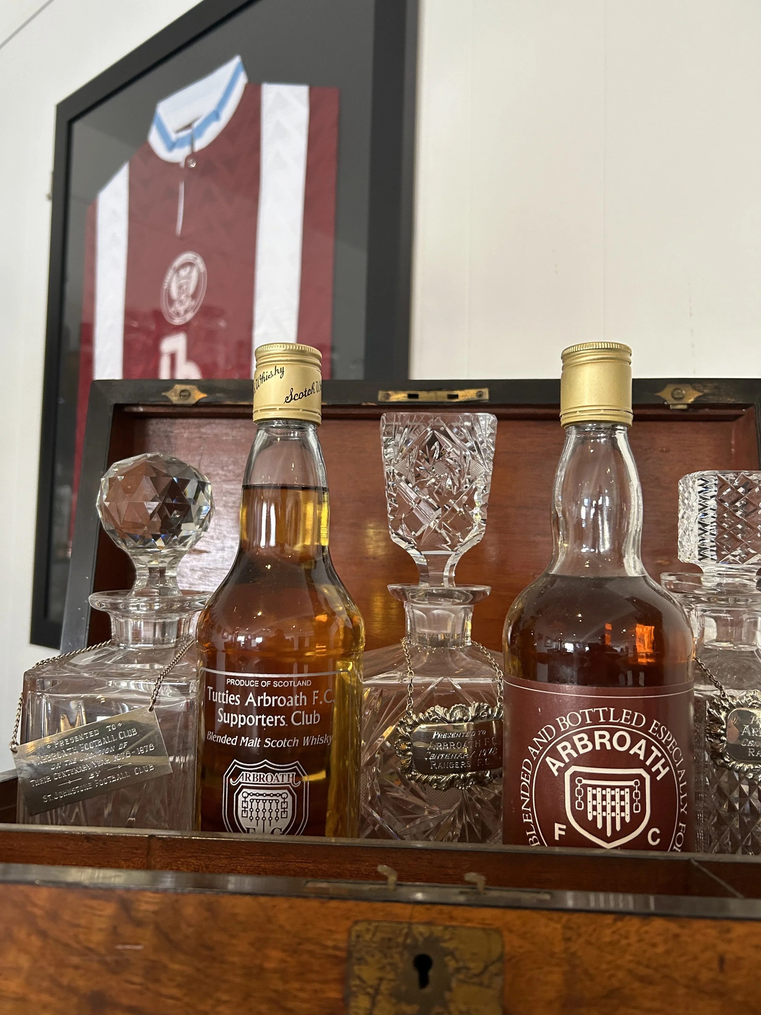 Glass bottles of Arbroath blended malt Scotch whisky on a wooden surface, with crystal glass decanters surrounding them, and a framed Juventus football jersey in the background.