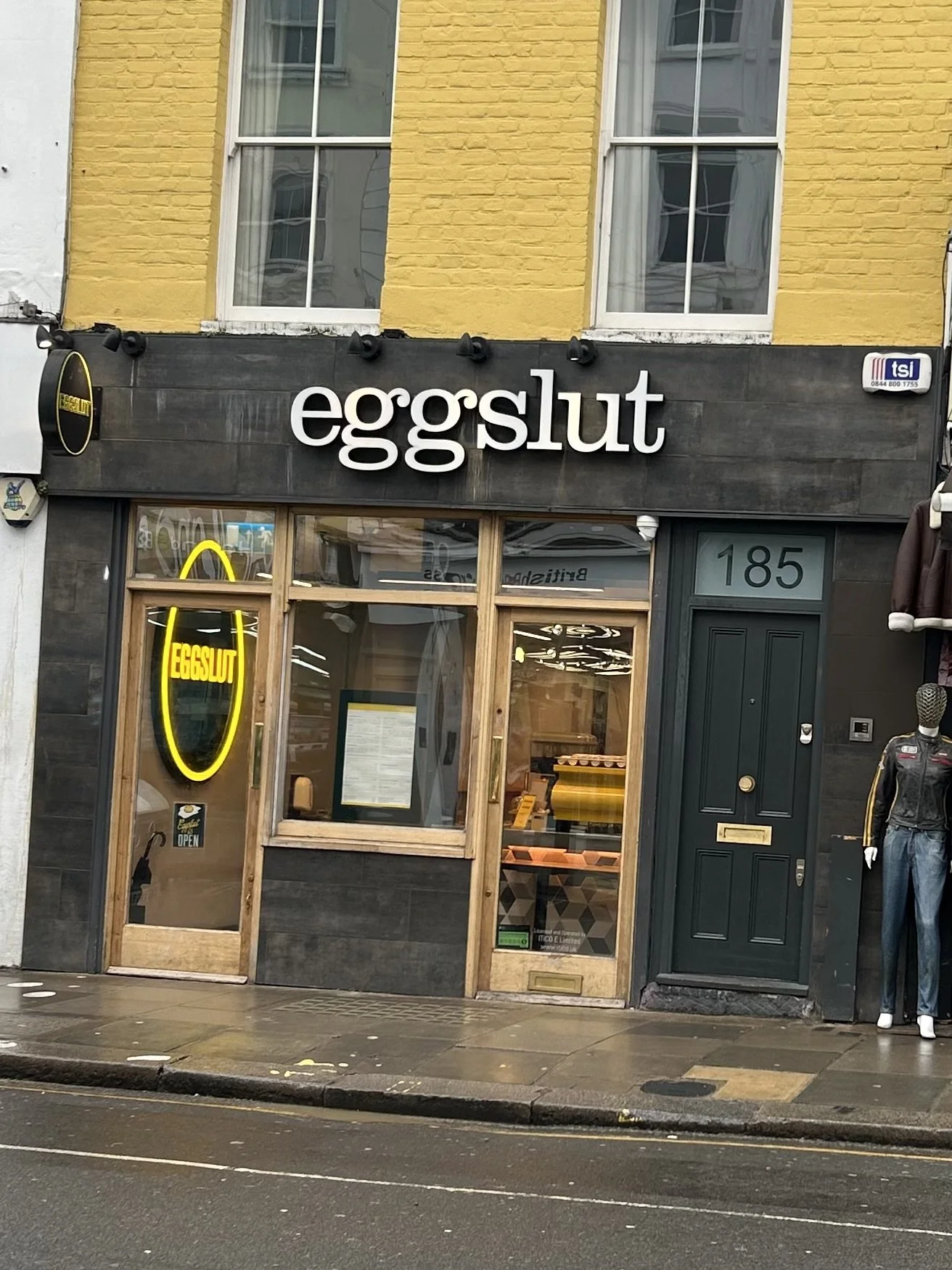  storefront named Eggslut with a black sign and large white letters, neon Eggslut sign on the door, mannequin wearing a jacket and jeans outside, and yellow painted building in the background.
