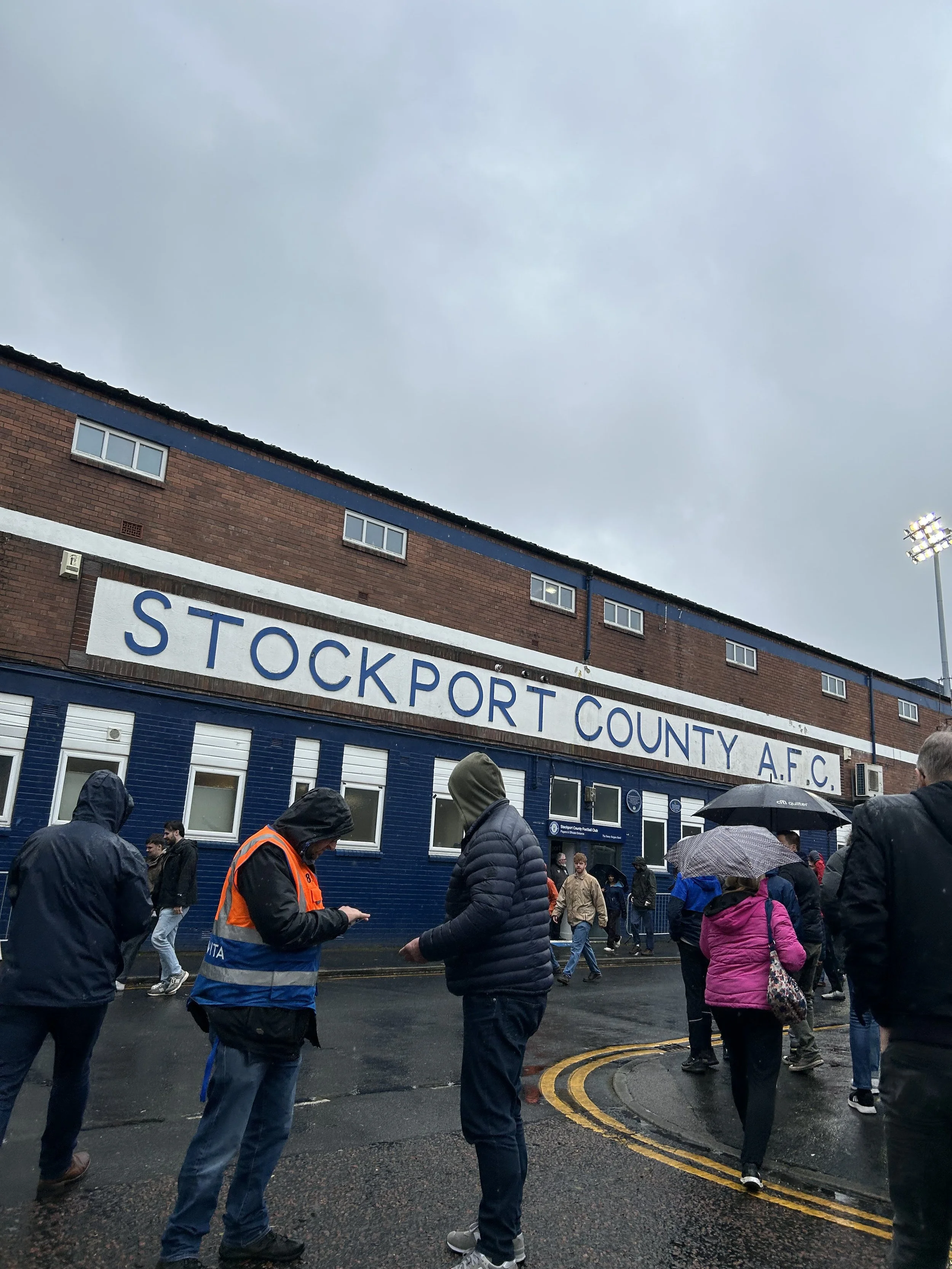 People walking outside in rainy weather near Stockport County A.F.C. stadium, with some holding umbrellas and wearing rain jackets.