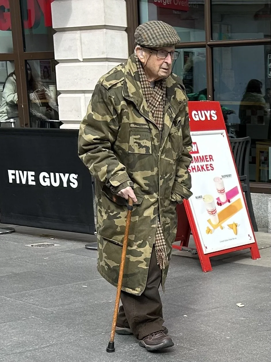 An elderly man in camouflage coat and flat cap walking with a cane outside a Five Guys restaurant, with a red menu board advertising summer shakes in the background.